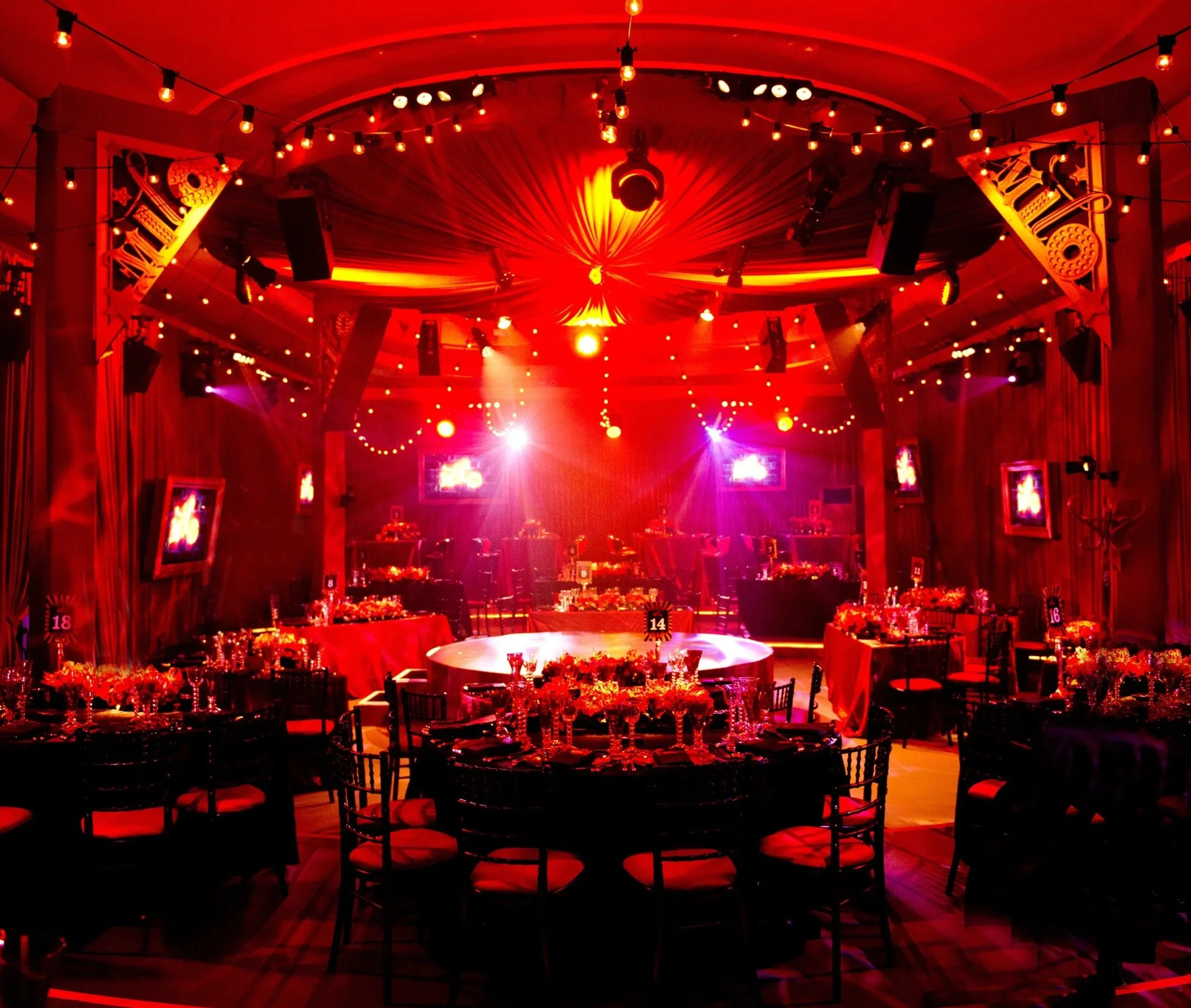 Red-lit banquet hall with round tables, decorated for an event, with floral centerpieces and glassware, illuminated by string lights and stage lighting.