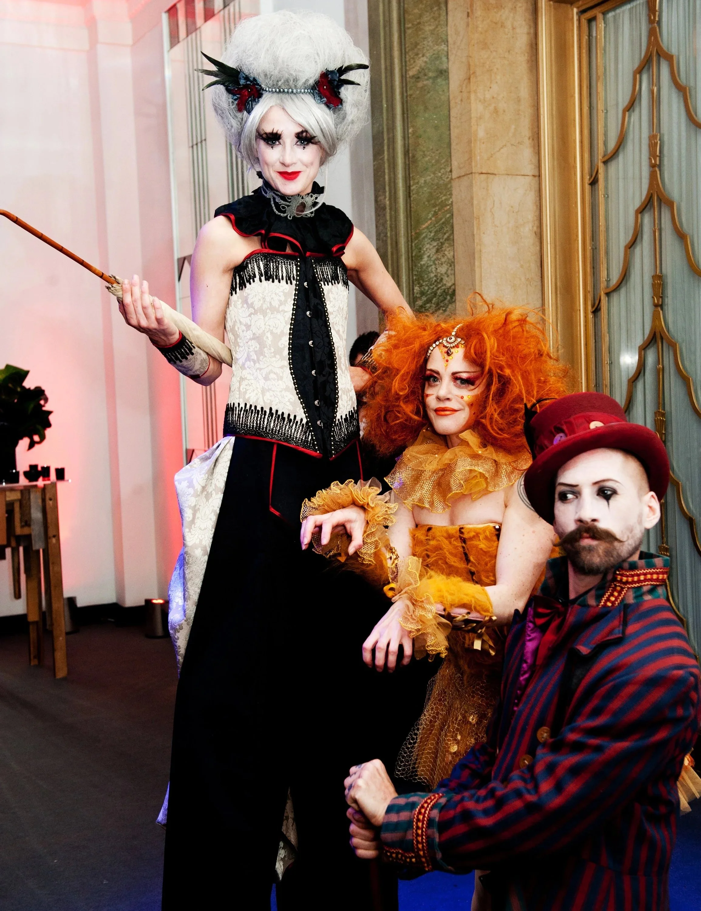 Two performers in elaborate costumes, one with a large white wig and floral headpiece, and the other with bright orange curly hair, makeup, and a gold ruffled dress, holding hands at a costume event.