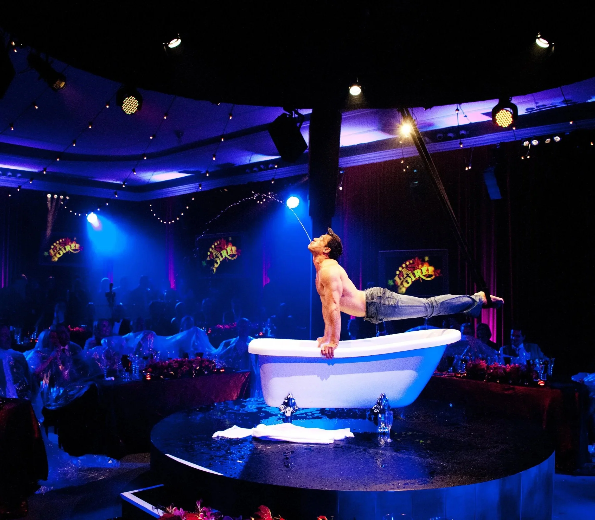 A male performer balancing on a bathtub in a circus or stage event, with theater lights and colorful decorations in the background.