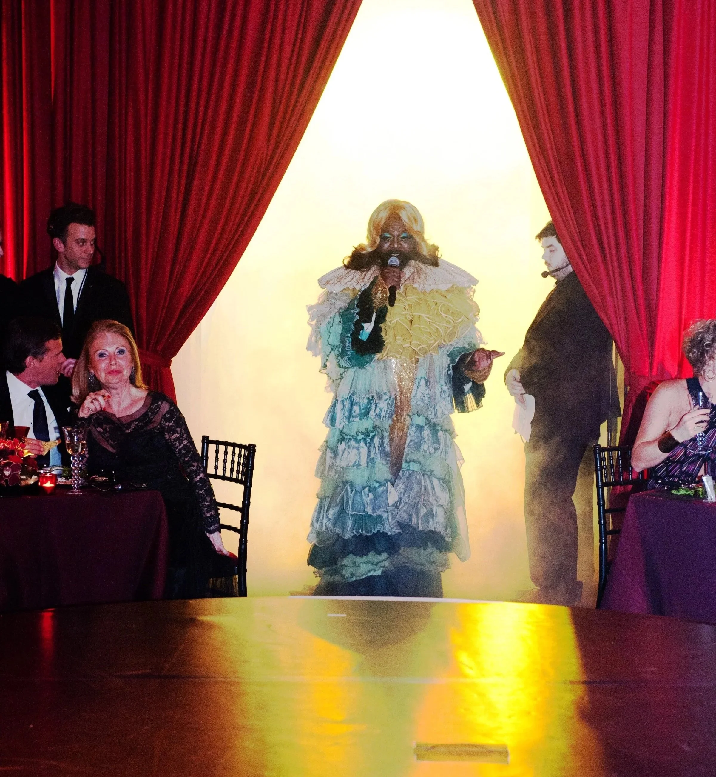 A performer in a colorful, elaborate costume with a blonde wig and makeup singing into a microphone on stage, surrounded by red curtains, with audience members visible.