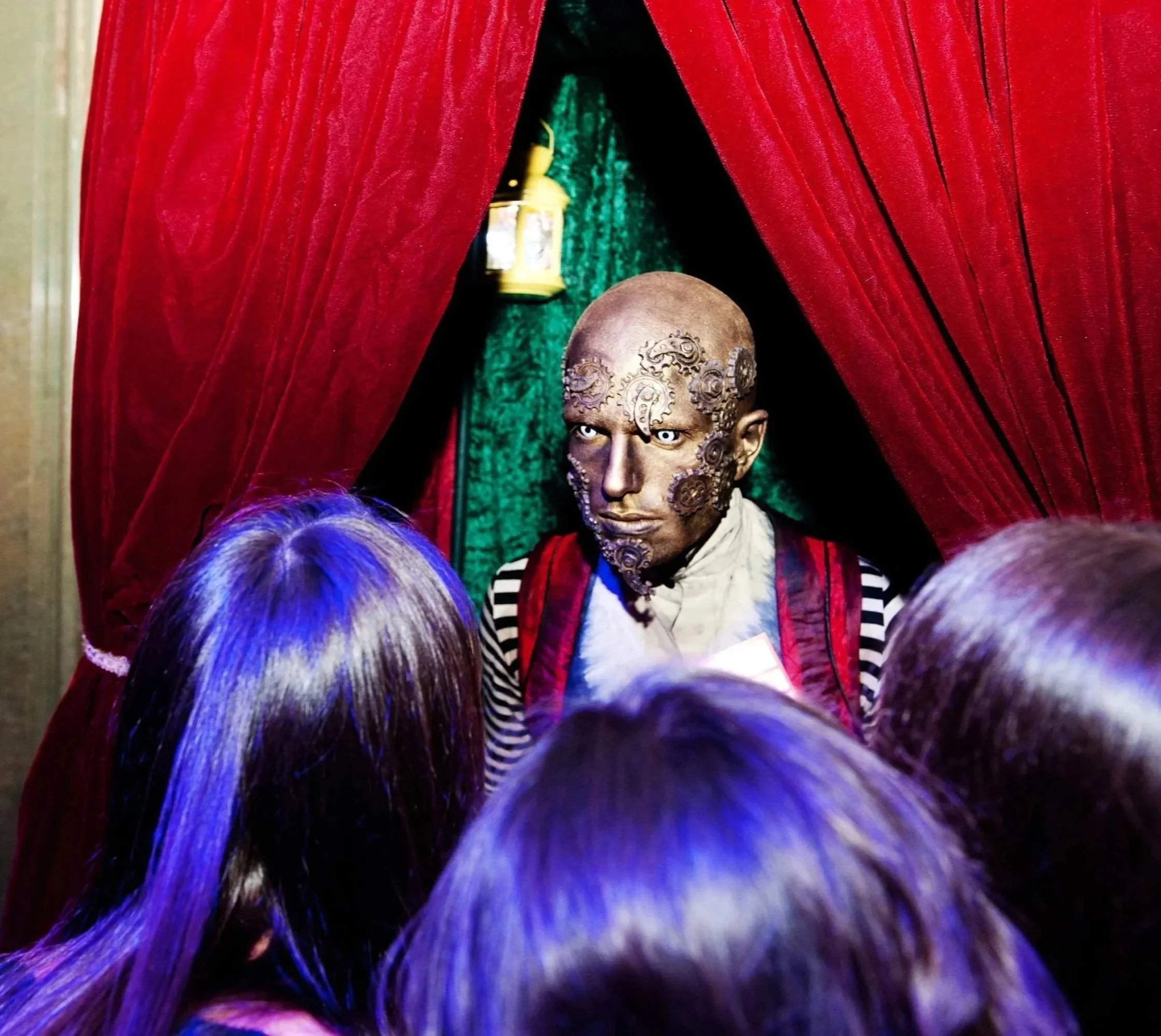 A person with intricate face tattoos, wearing a striped shirt and a red vest, peering from behind red velvet curtains, with purple hair and a cloth covering part of their face.