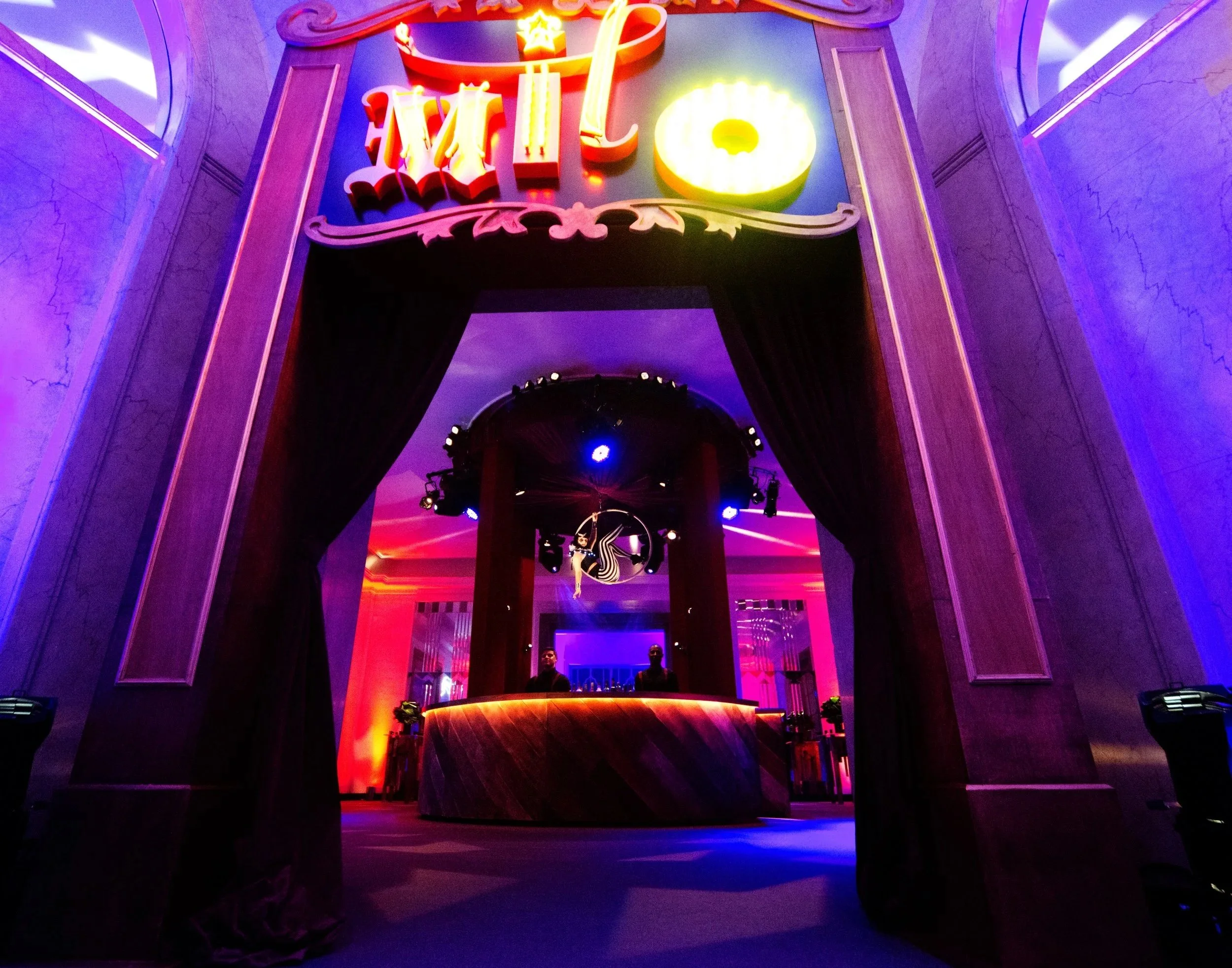 Colorful nightclub bar with neon lights, a circular bar counter, two staff members behind the bar, and a DJ at the turntables in the background.