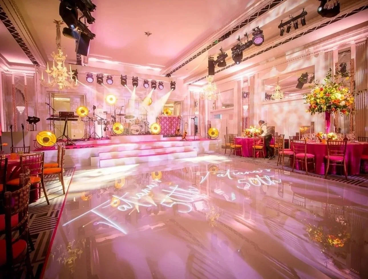 Elegant event hall decorated with pink lighting, tables with pink tablecloths, floral centerpieces, a stage with musical instruments, and reflective shiny flooring with event signage projected onto it.