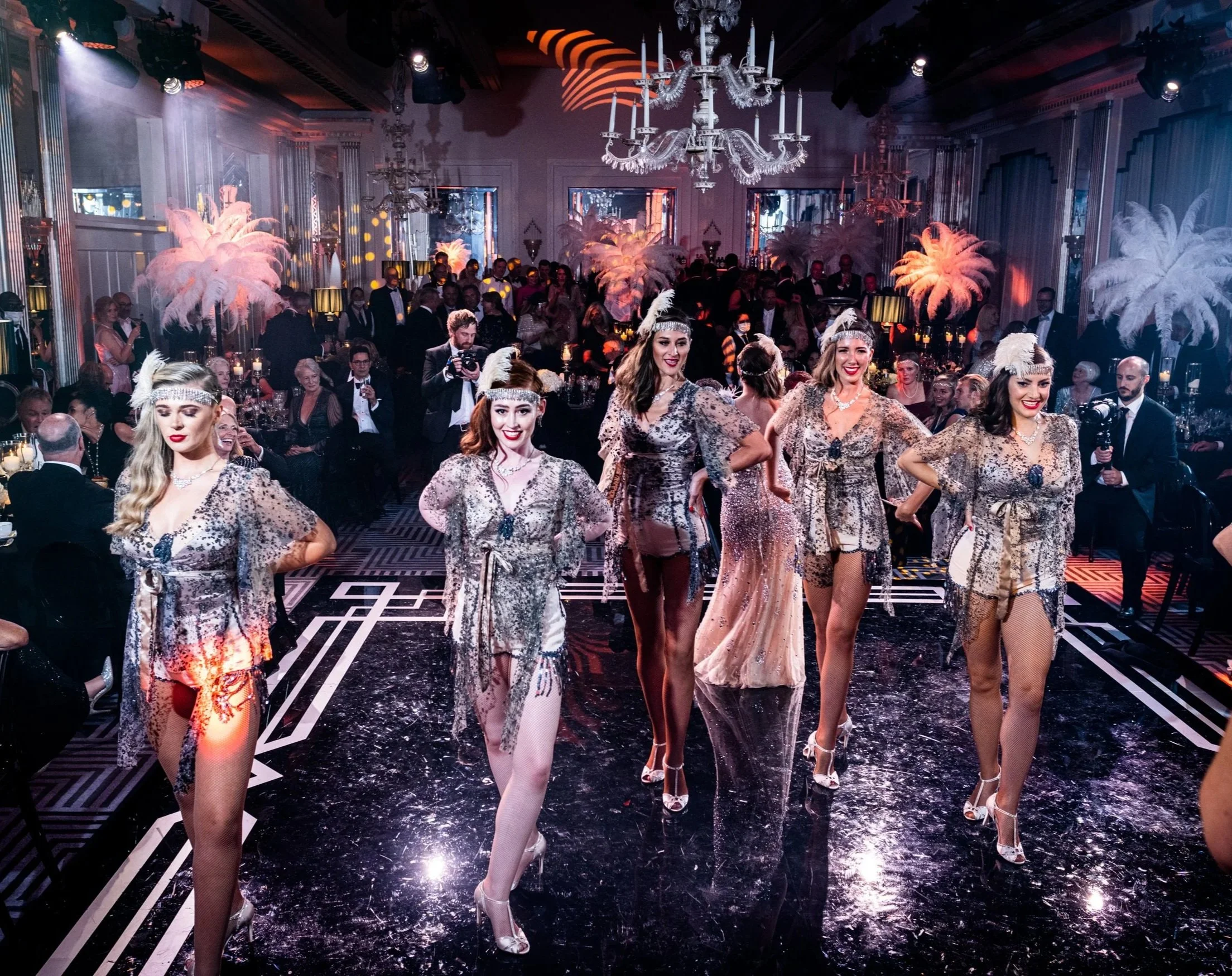 A group of women dressed in elegant, matching animal print dresses with feathers and feathered headbands, walking confidently at a glamorous indoor event with chandeliers, feather decorations, and an audience seated at tables.