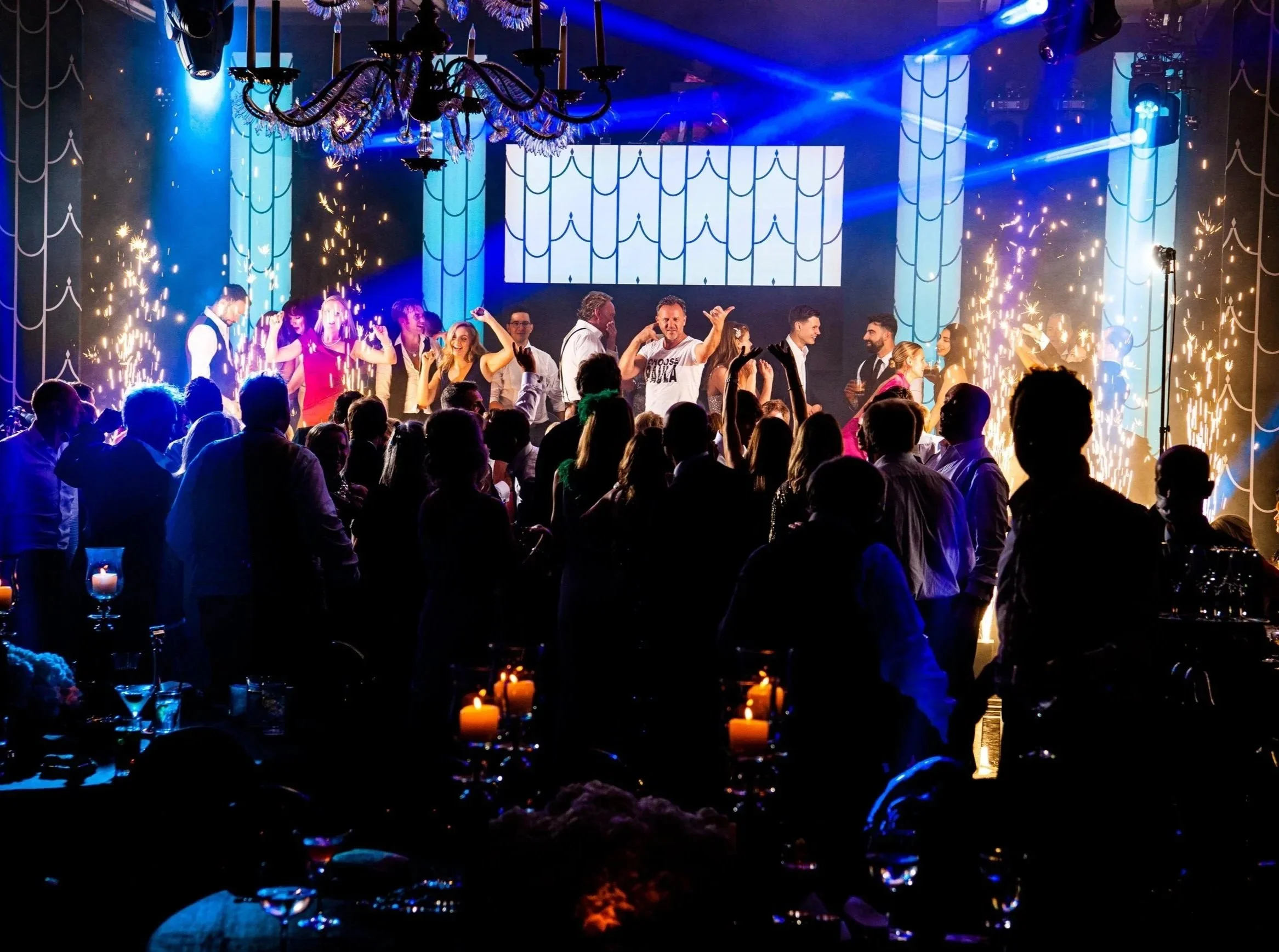 A lively party scene with a group of people dancing and celebrating on a stage illuminated by colorful lights and fireworks, with a chandelier hanging from the ceiling and candles on tables in the foreground.