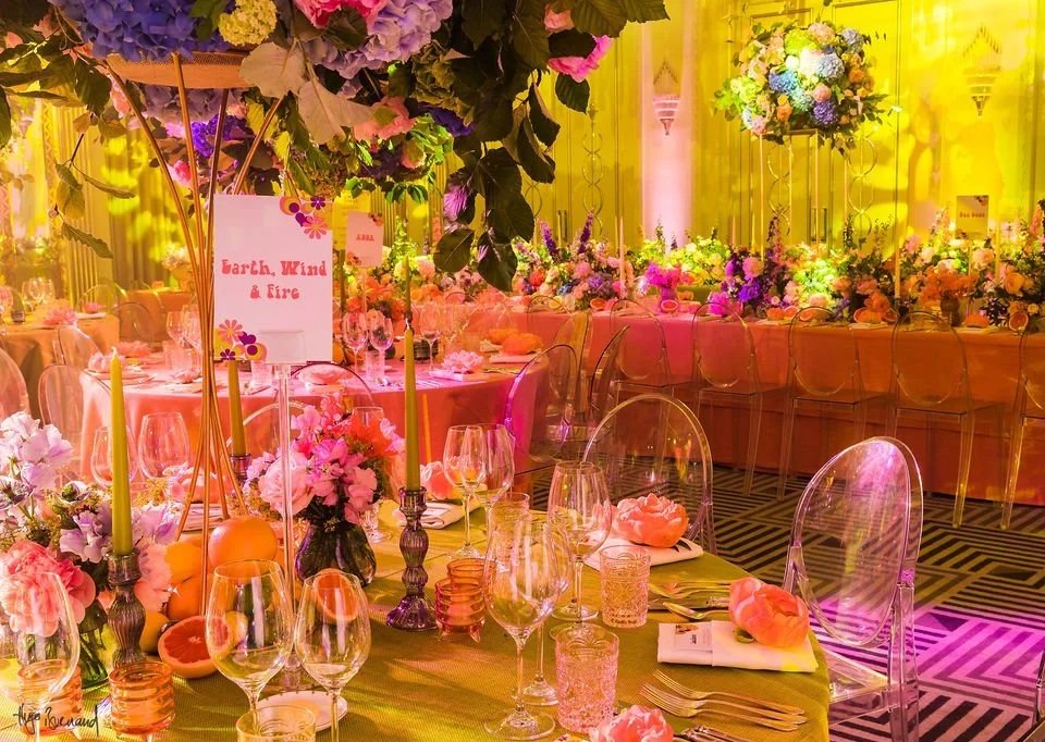Elegant Claridge's decorated for a 70's Bat Mitzvah with floral arrangements, candles, and glassware on tables, decorated in pink, purple, and yellow lighting.