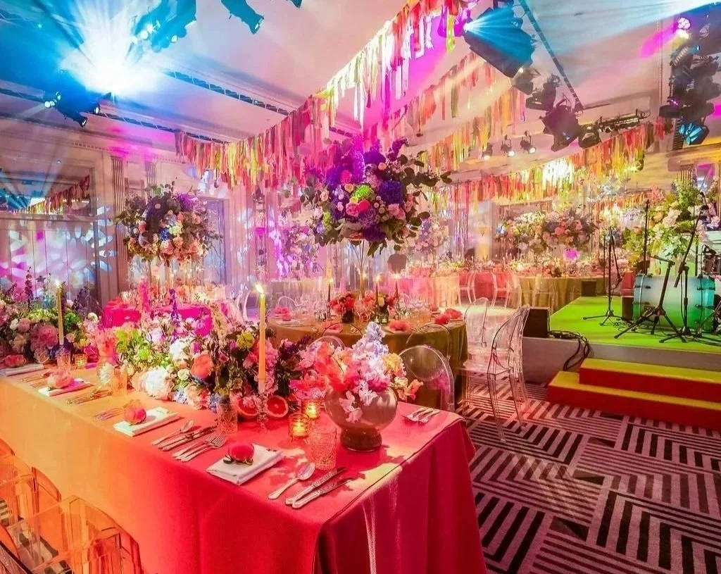 A colorful Claridge's space decorated with flowers, hanging strands of fabric, and vibrant lighting. There are long tables with floral centerpieces and place settings, and a small stage area with musical instruments.