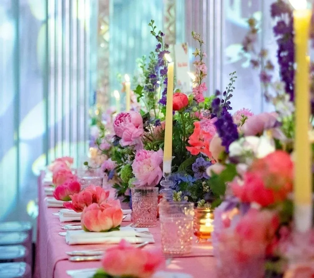 A table decorated with pink and purple flowers, pink and peach peonies, and candles, set for a celebration or event in a bright space.