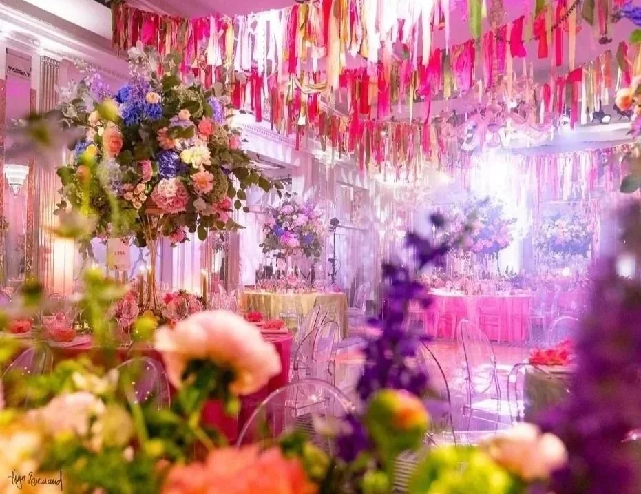 Decorated event hall with pink, purple, and gold ribbons hanging from the ceiling, floral arrangements with roses and other flowers, transparent chairs, and pink tablecloths.