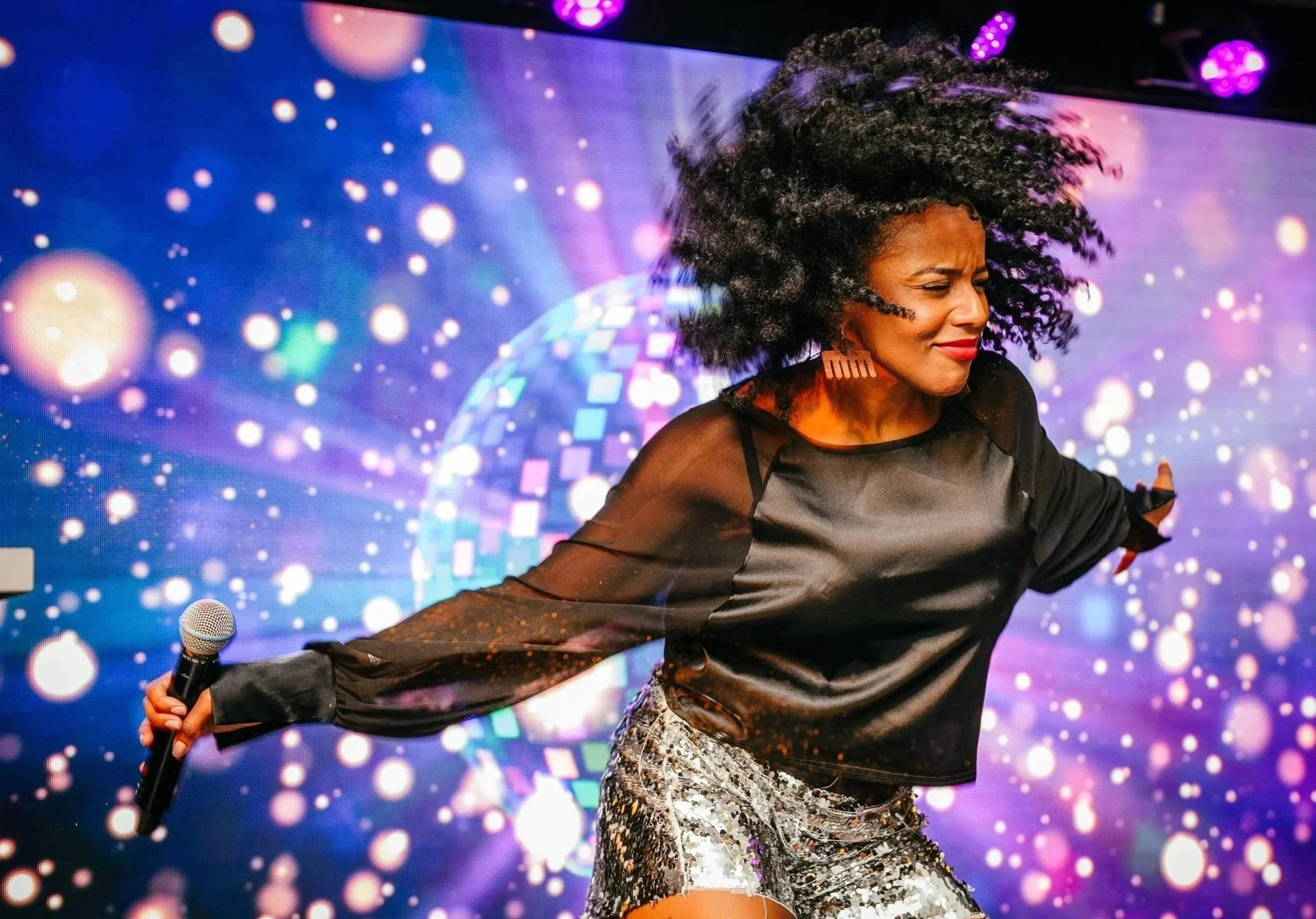 A woman with curly hair wearing a shiny black top and patterned shorts on stage with a colorful, dotted background, holding a microphone in her right hand.