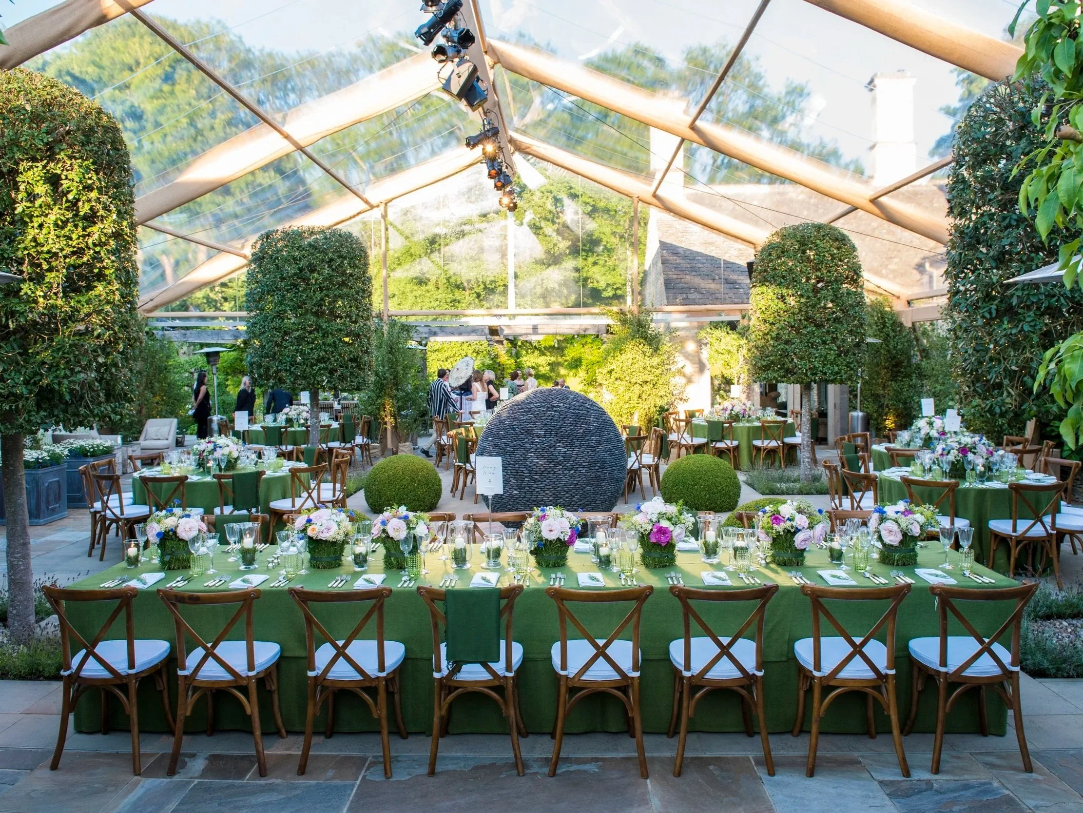 Wedding reception setup with a long green-draped table, floral centerpieces, and chairs on a stone patio under a clear tent with visitors and greenery.