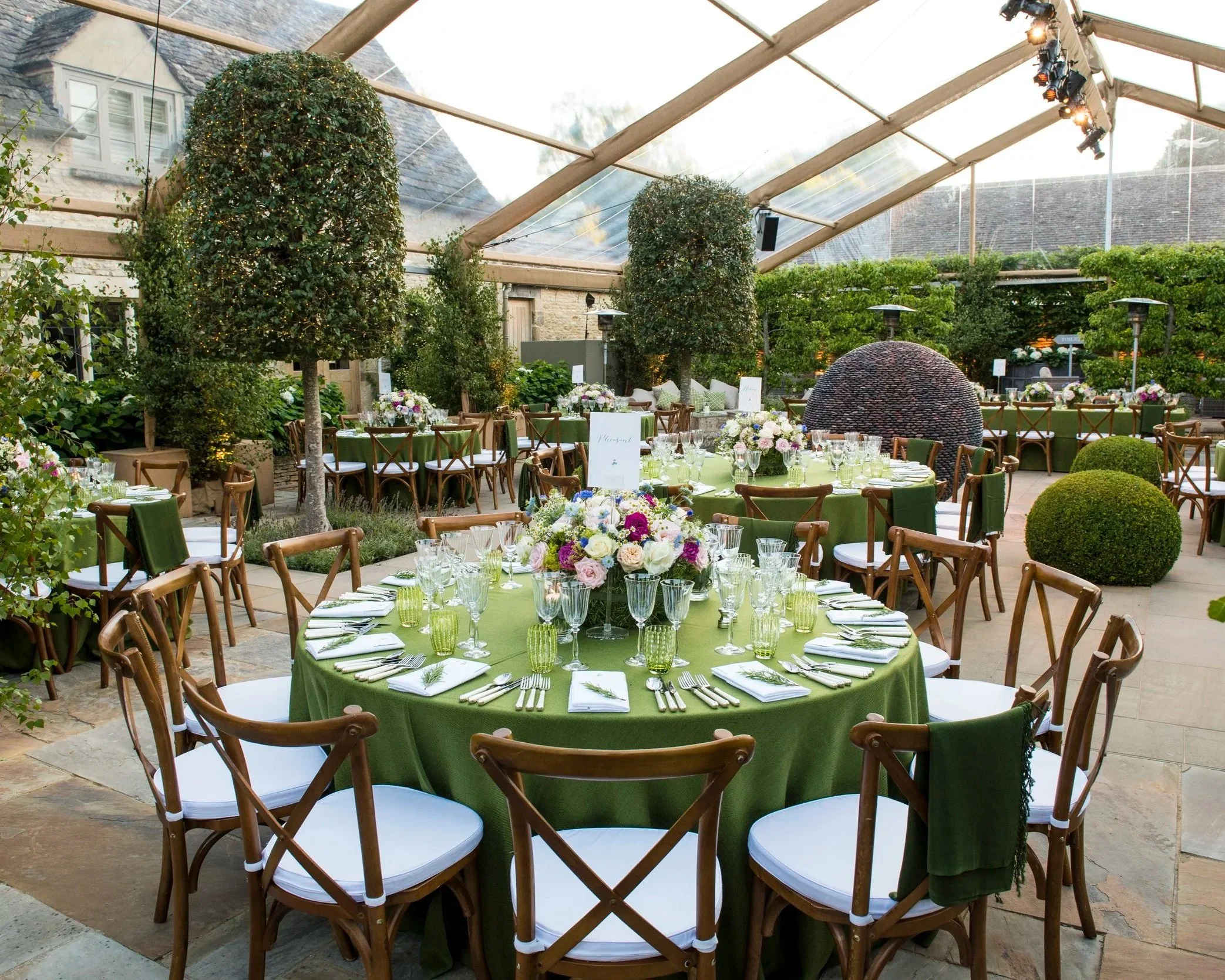 Indoor event space with decorated tables and greenery, set for a banquet with floral centerpieces and green tablecloths.