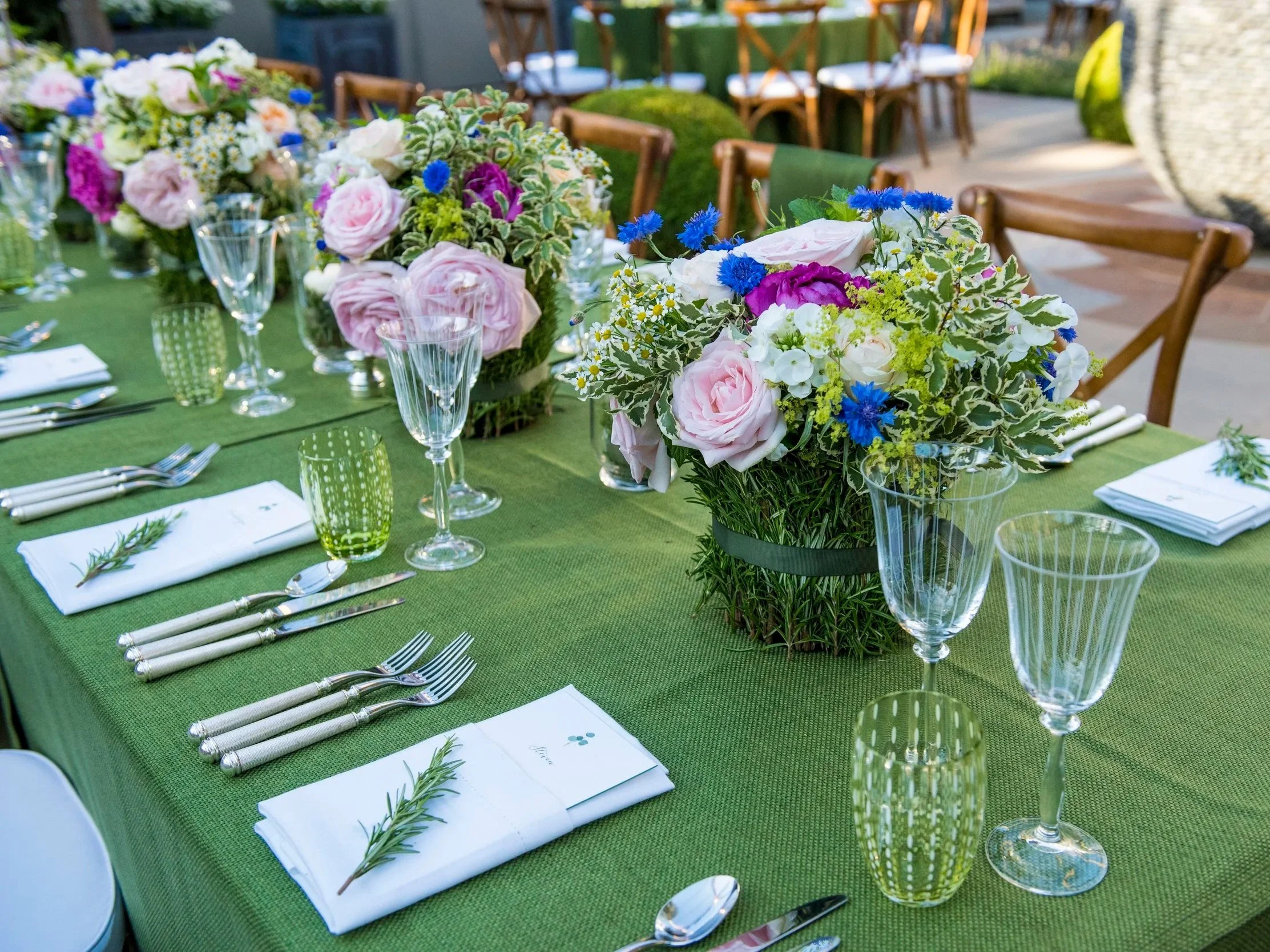 A table set for a formal outdoor meal with floral centerpieces featuring pink, purple, white, and blue flowers, green foliage, clear wine glasses, green patterned drinking glasses, white napkins with herbs, and silverware on a green tablecloth.