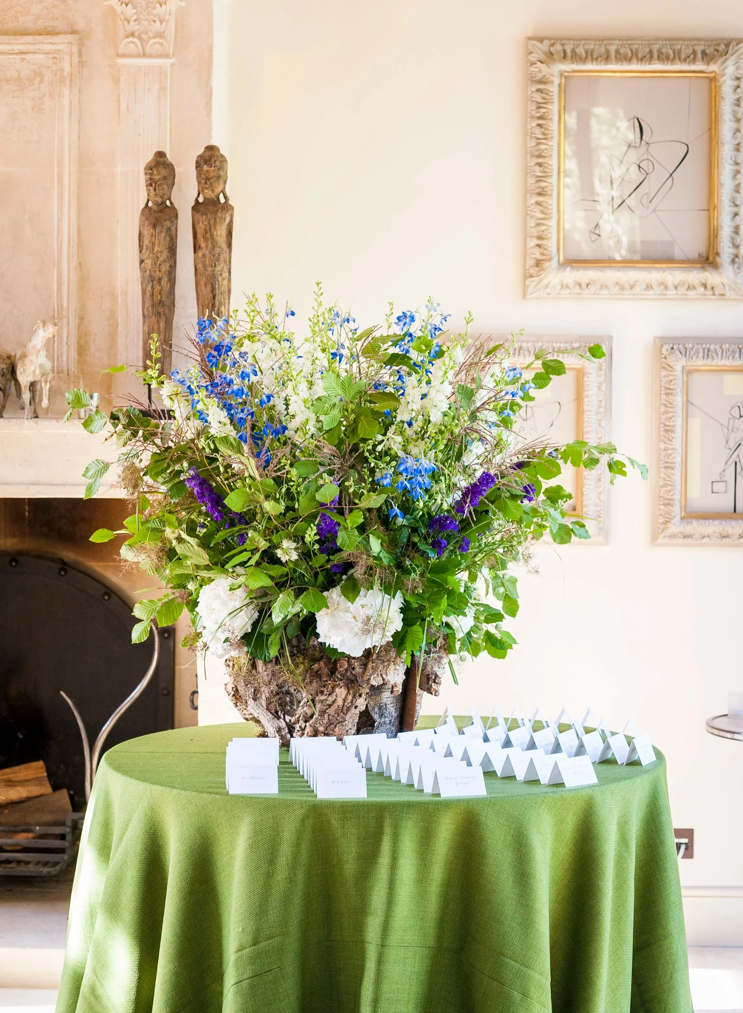 A large flower arrangement with white, blue, purple, and green flowers on a round table covered with a green tablecloth. Display cards are lined up in front of the arrangement. Framed artwork hangs on a cream wall in the background.