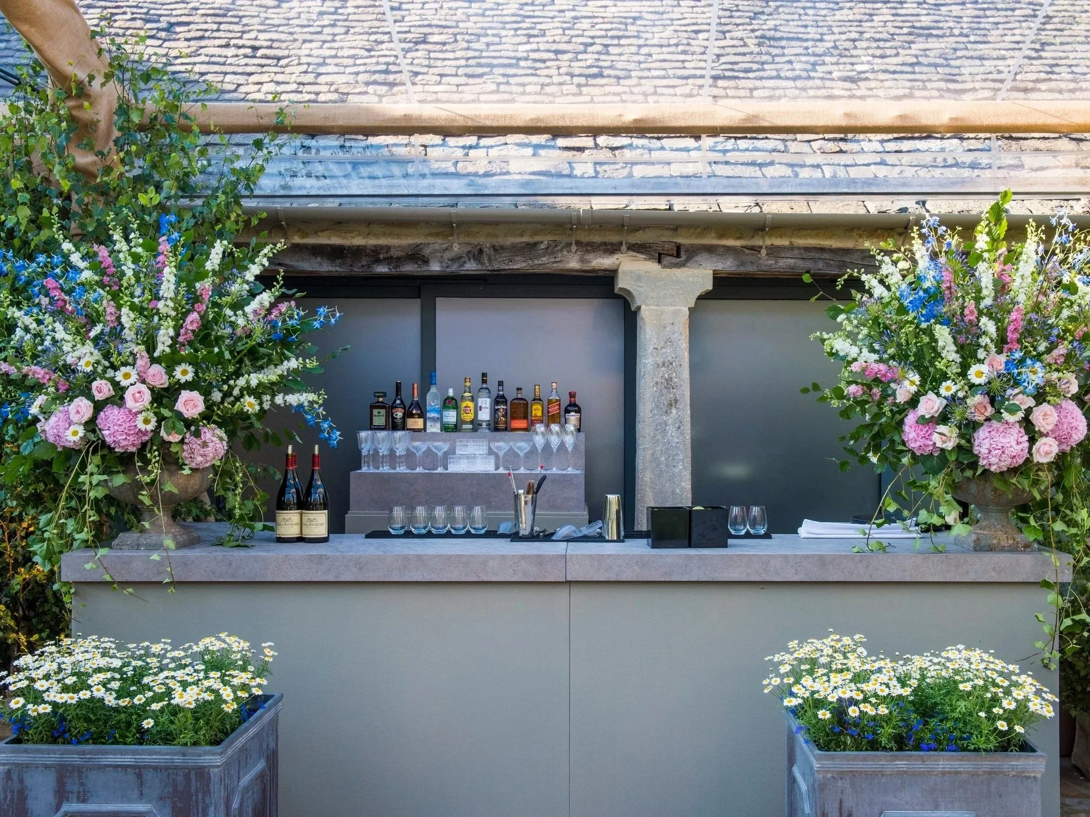 Outdoor bar setup with bottles of alcohol and glassware, flanked by large colorful flower arrangements on planters, against a building with brick and stone exterior.