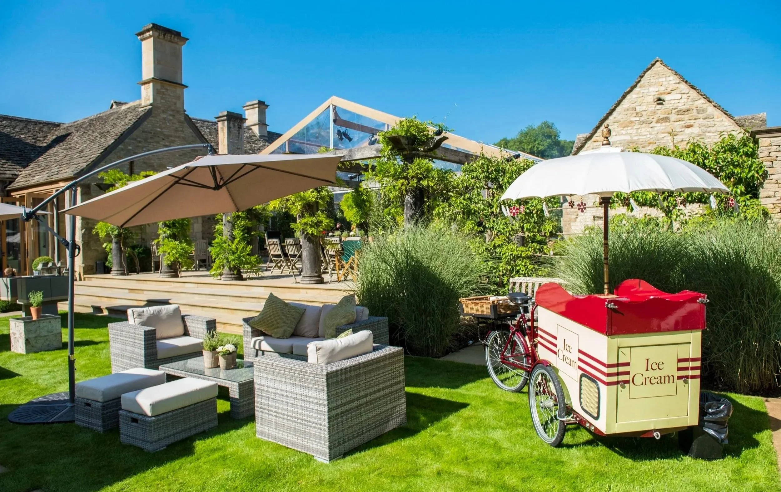 A backyard patio with wicker furniture, umbrellas, and lush green grass. There is an ice cream cart with a red bicycle attached, set against a backdrop of stone houses and vibrant plants under a clear blue sky.