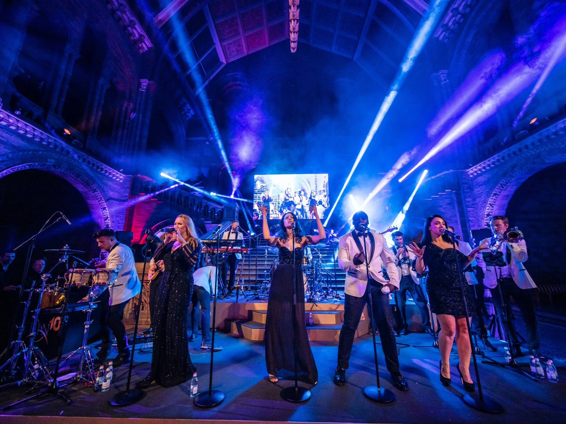 A live musical performance features a group of singers and musicians on an illuminated stage with blue and purple lighting at The Natural History Museum