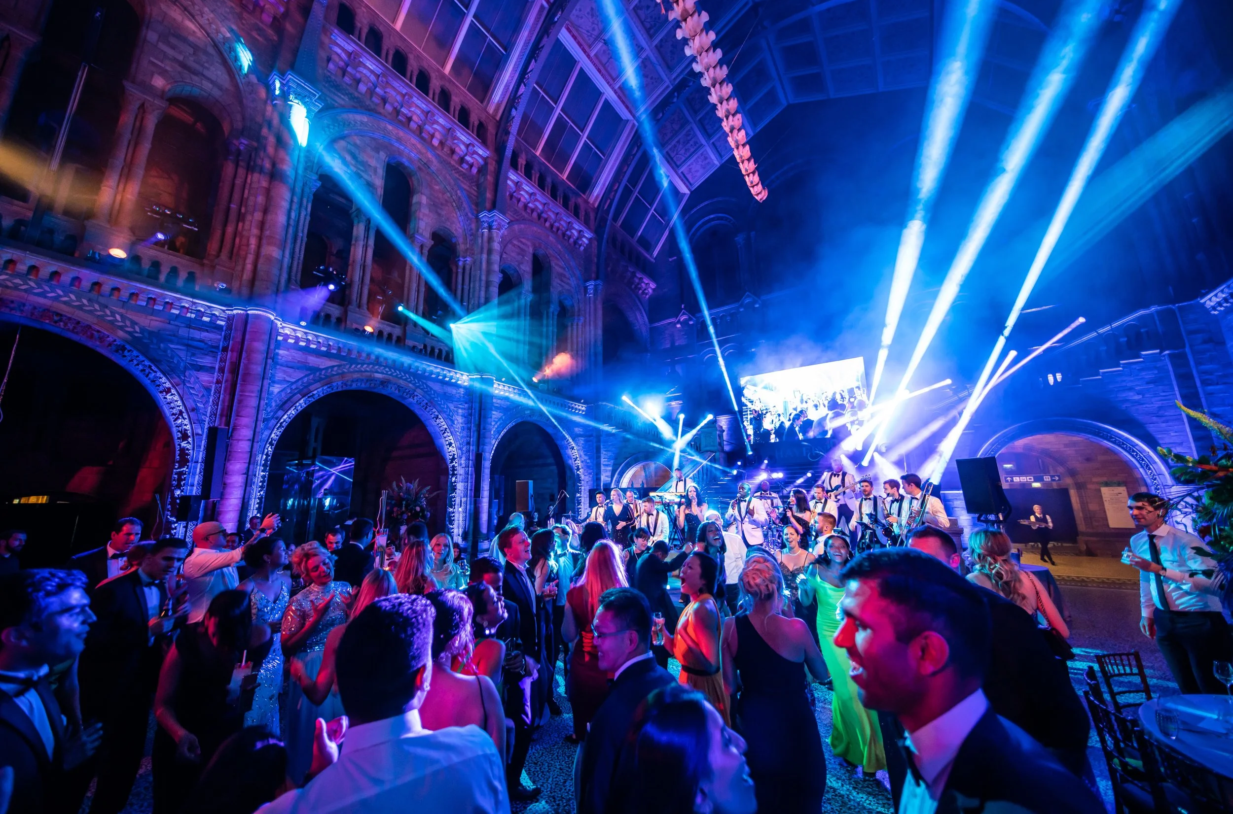 A lively party or concert inside a grand, architecturally detailed building with high, arched ceilings and large windows. The space is illuminated with colorful, dynamic stage lighting, and musicians are performing at The Natural History Museum