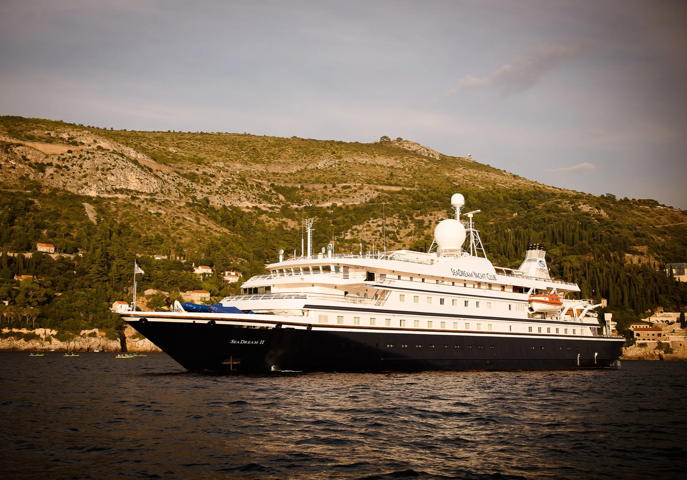 A large white yacht named Sea Dream II docked in a bay with a hillside covered in trees and small buildings in the background.