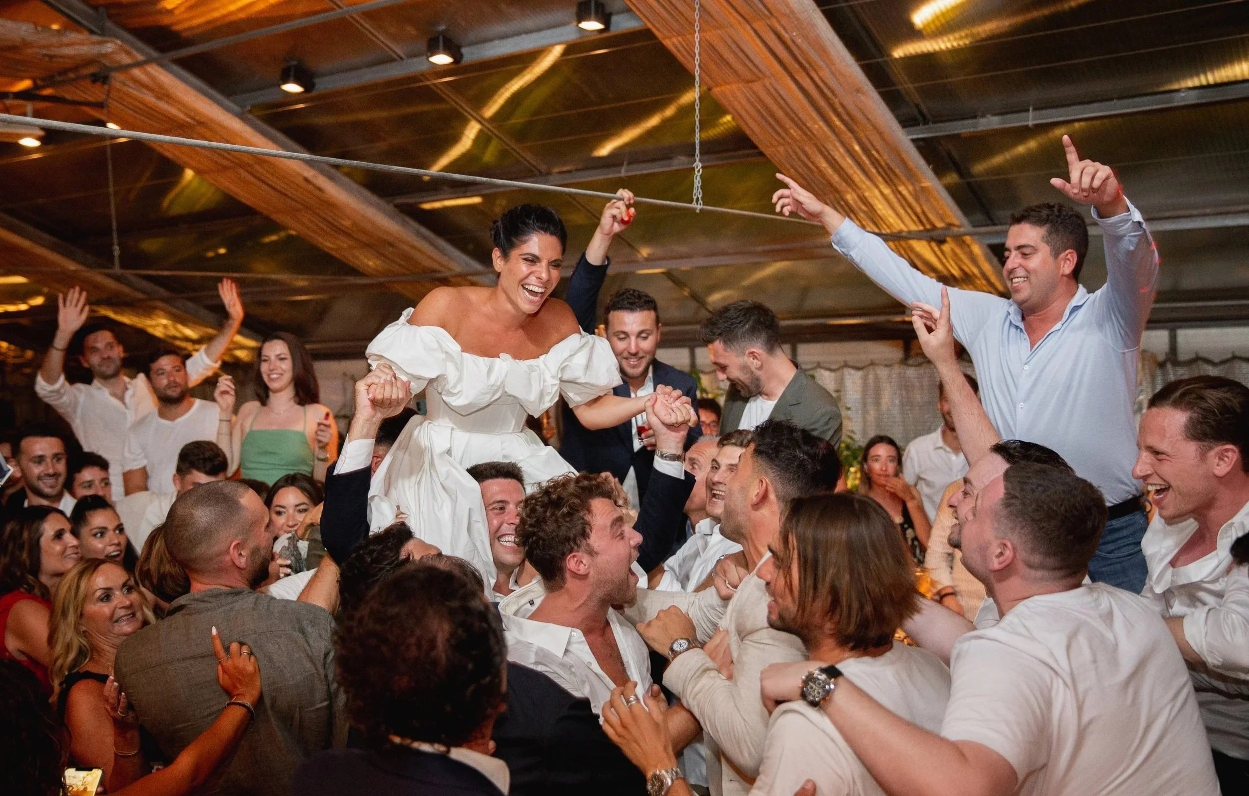 A lively celebration with a large crowd of diverse people, some smiling and cheering, as a woman in a white dress is lifted above the crowd, smiling joyfully. The setting appears to be indoors with a ceiling of wooden panels and warm lighting.