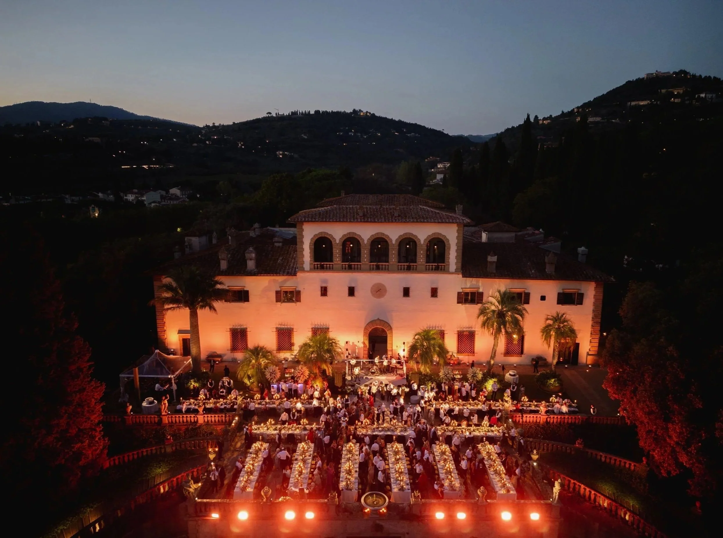 An evening outdoor wedding reception at a historic mansion called Villa Palmieri in Florence, with guests seated at long tables adorned with candles and floral centerpieces, overlooking a scenic mountain landscape.