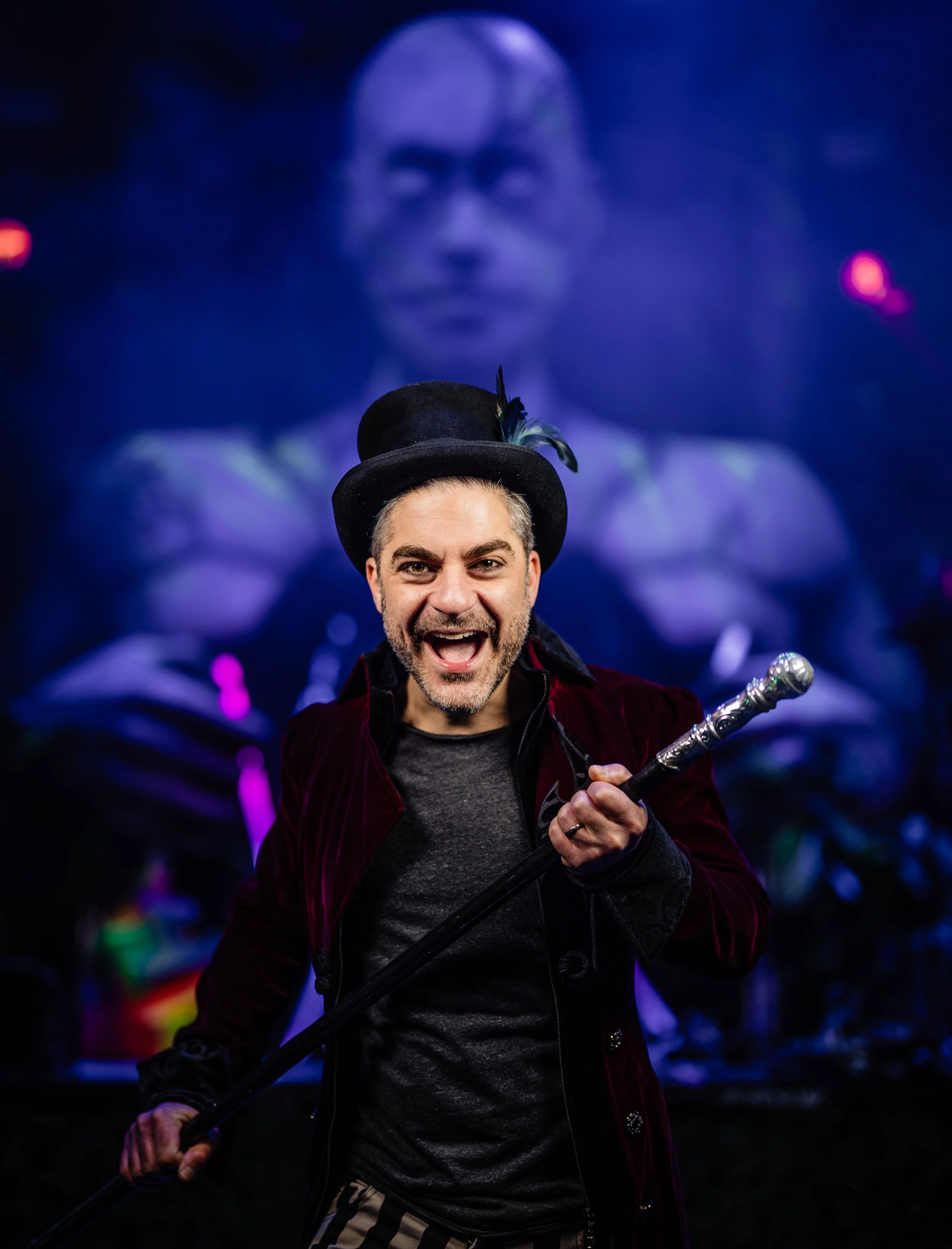 Man wearing a black hat with a feather, smiling, holding a staff, standing in a dark setting with a large blurred projection of a person's face in the background.
