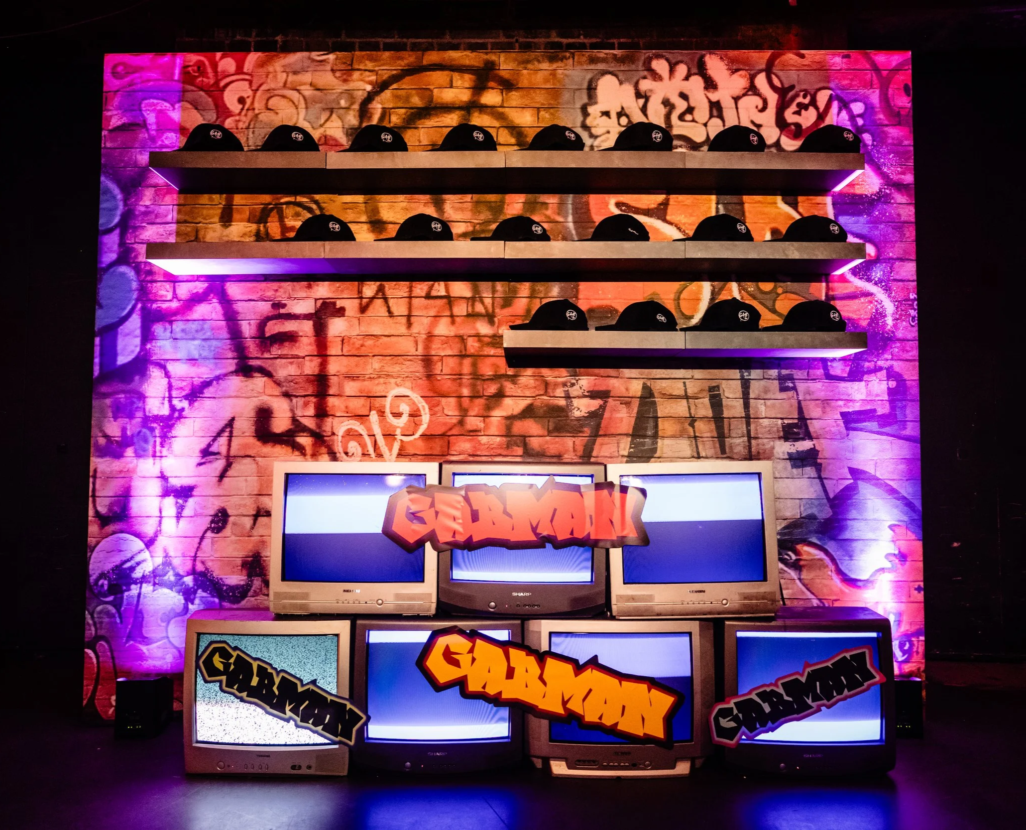 This image shows a colorful display of vintage CRT televisions stacked on top of each other against a graffiti wall with bright pink and purple lights. The screens show various graffiti-themed graphics and text.