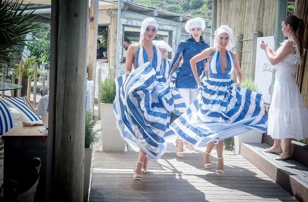 Three women in blue and white striped dresses and white head coverings dancing on a wooden deck, with a woman in a white dress taking a photo nearby, in an outdoor setting with bamboo fencing and greenery.