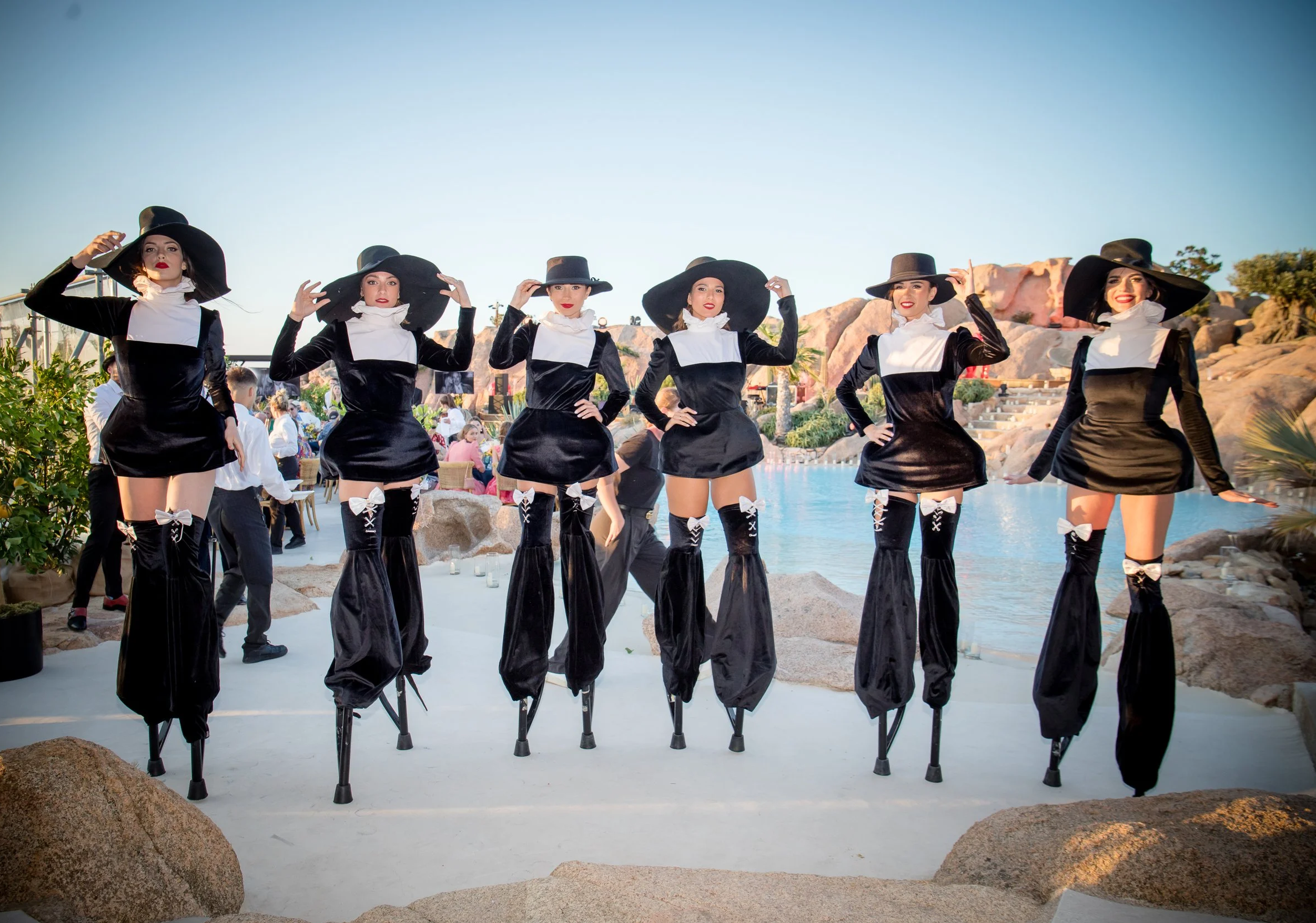 Six women dressed as witches, standing with crutches reversed on their hands, outdoors by a pool with rocks and plants, in front of a rocky landscape, under a clear sky.