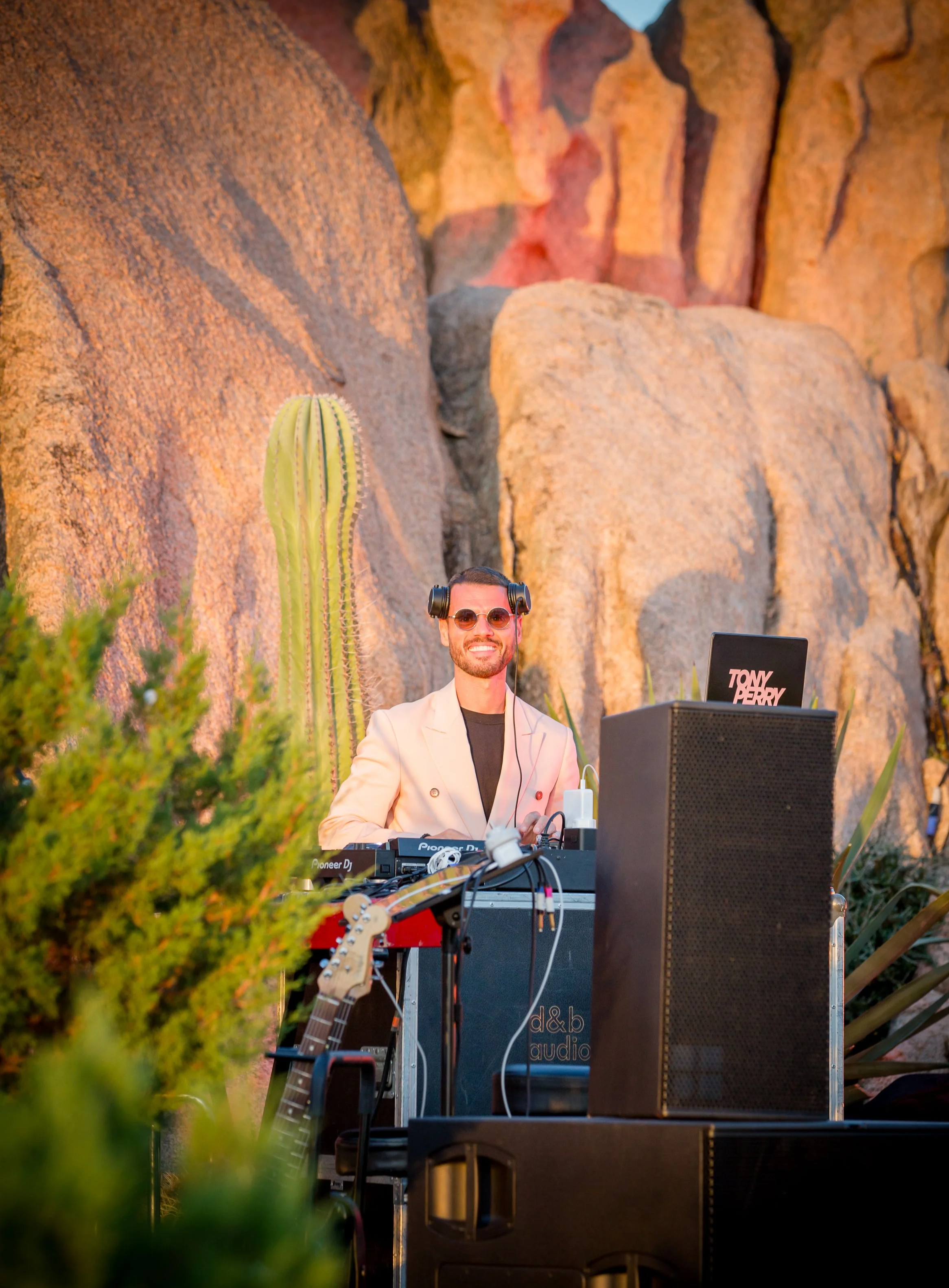 A man with sunglasses and a light blazer is DJing at an outdoor event, surrounded by large rocks and desert plants, including a saguaro cactus, during sunset.