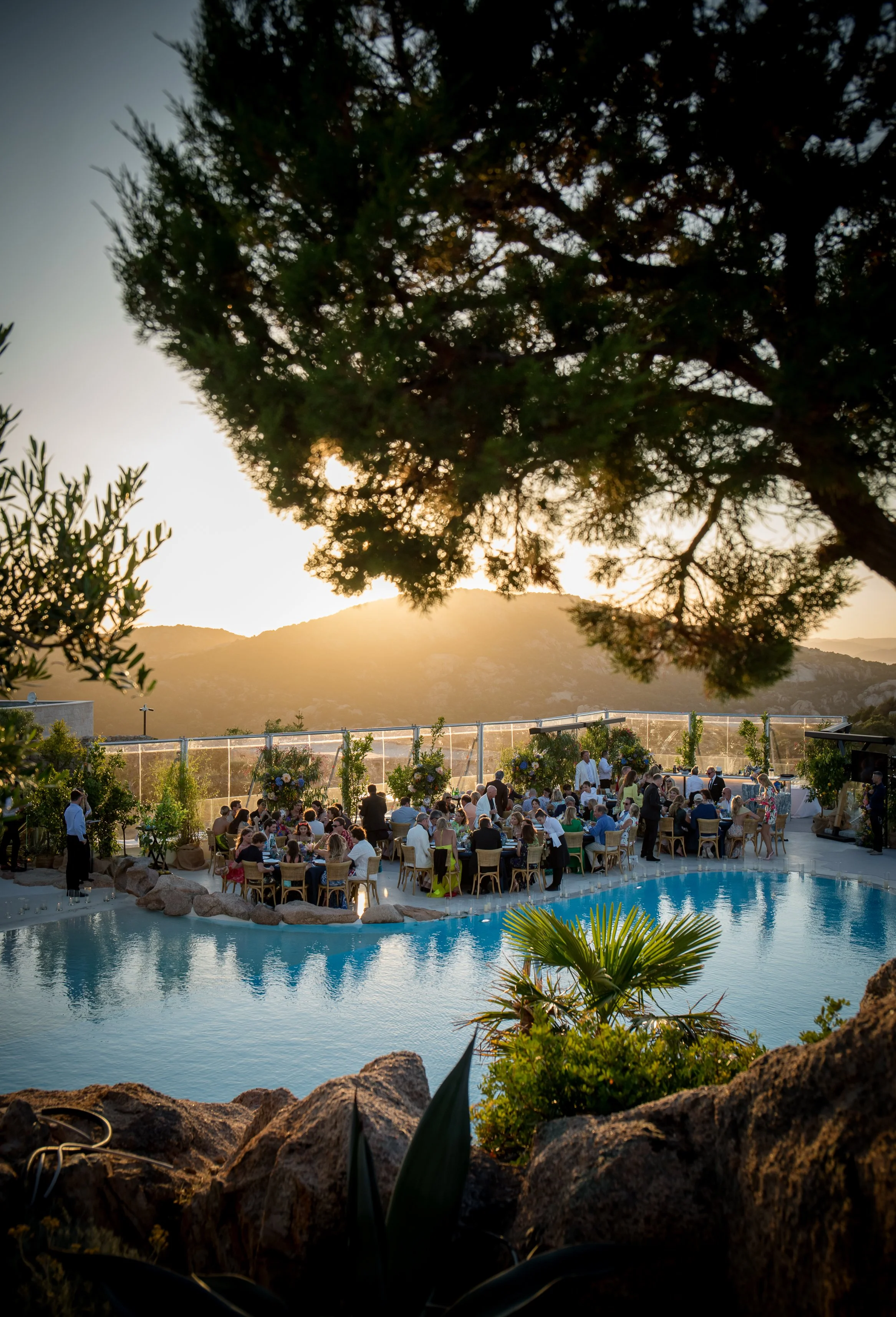 An outdoor event at sunset with people gathered around tables near a swimming pool, surrounded by trees and mountains in the background.