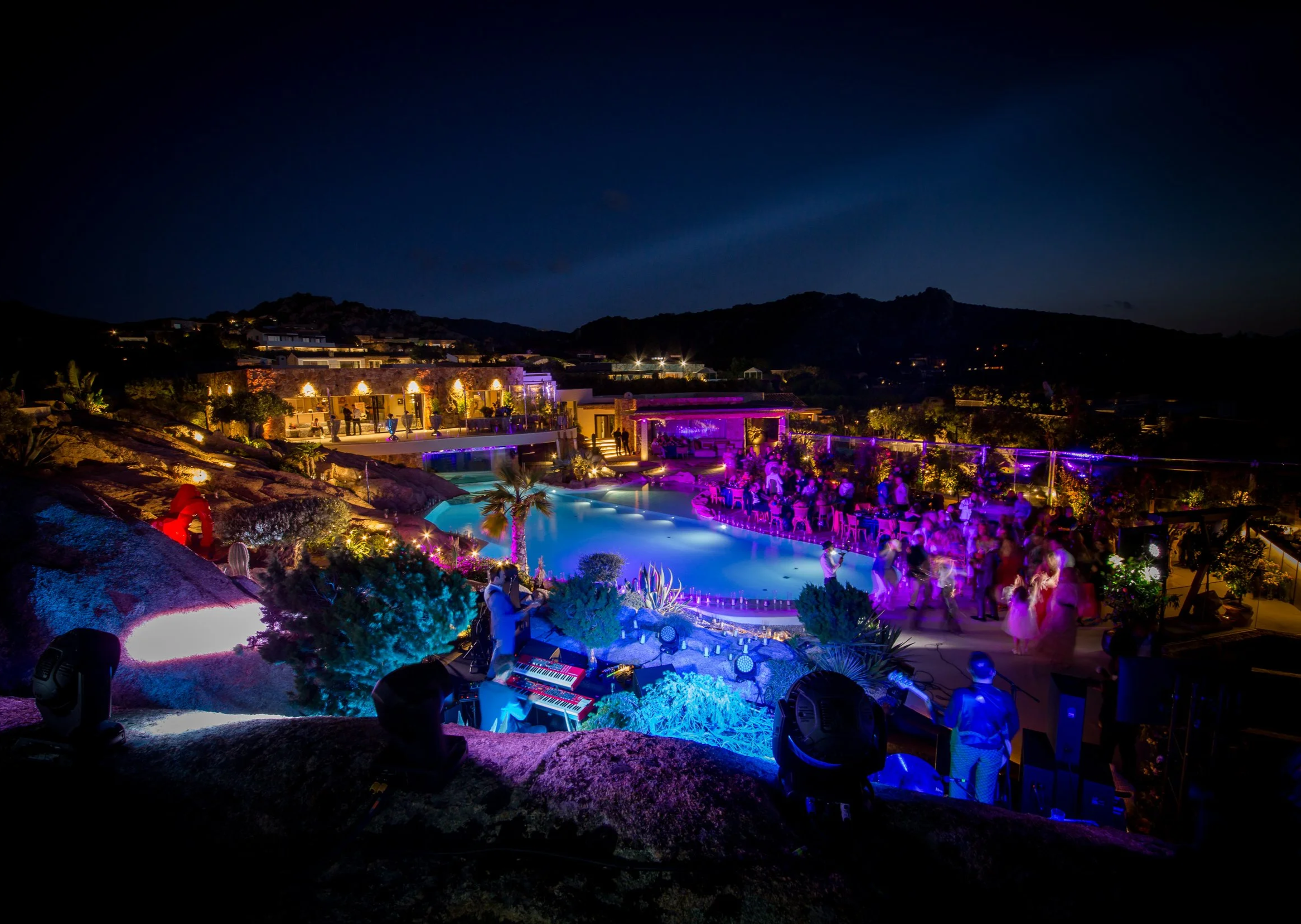Night scene of an outdoor party with colorful lighting, a pool, and a view of mountains in the background.