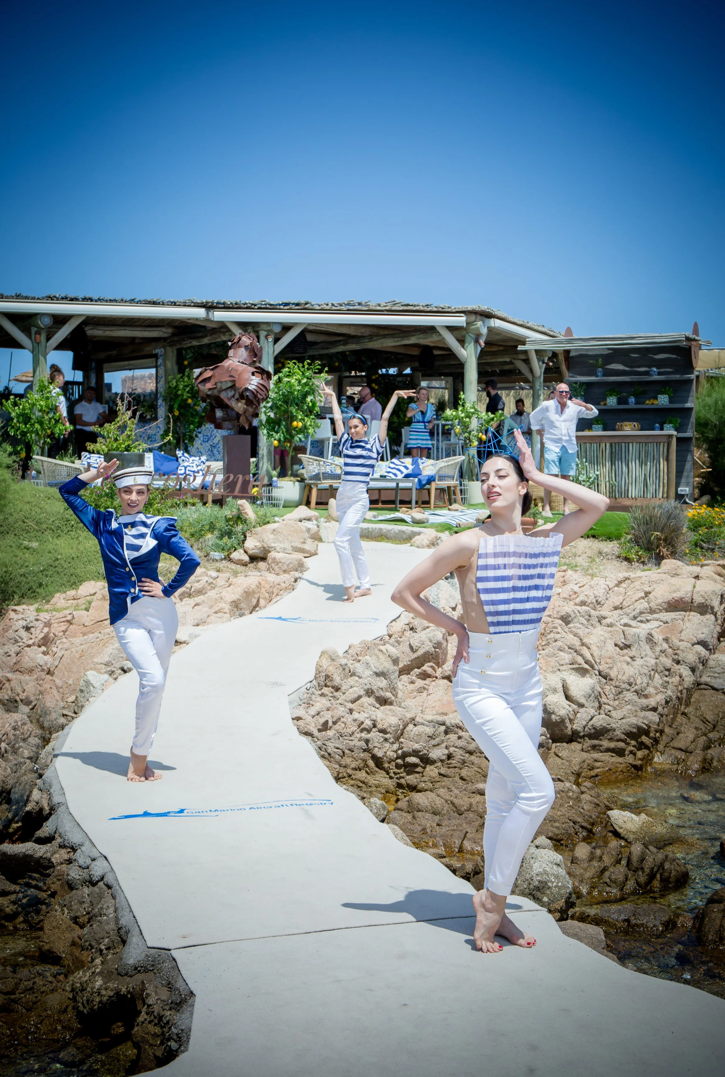 People dressed in nautical costumes, standing and posing on a white pathway with rocks on either side, near a building with outdoor seating and greenery, under a clear blue sky.