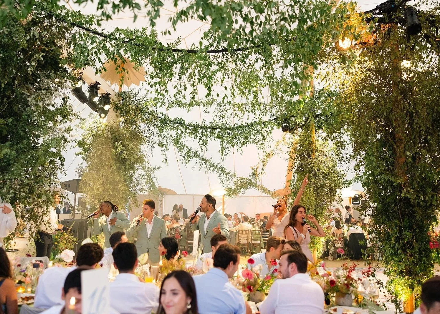 People singing and dancing at Hertfordshire private residents wedding reception inside a decorated venue with greenery and string lights.