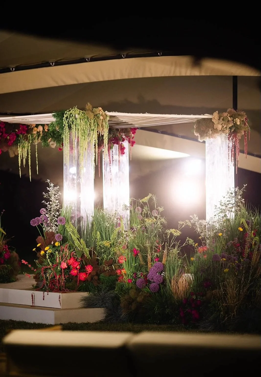 Lit up Chuppah floral arrangement with hanging curtains, illuminated by bright lights, featuring various colorful flowers and greenery