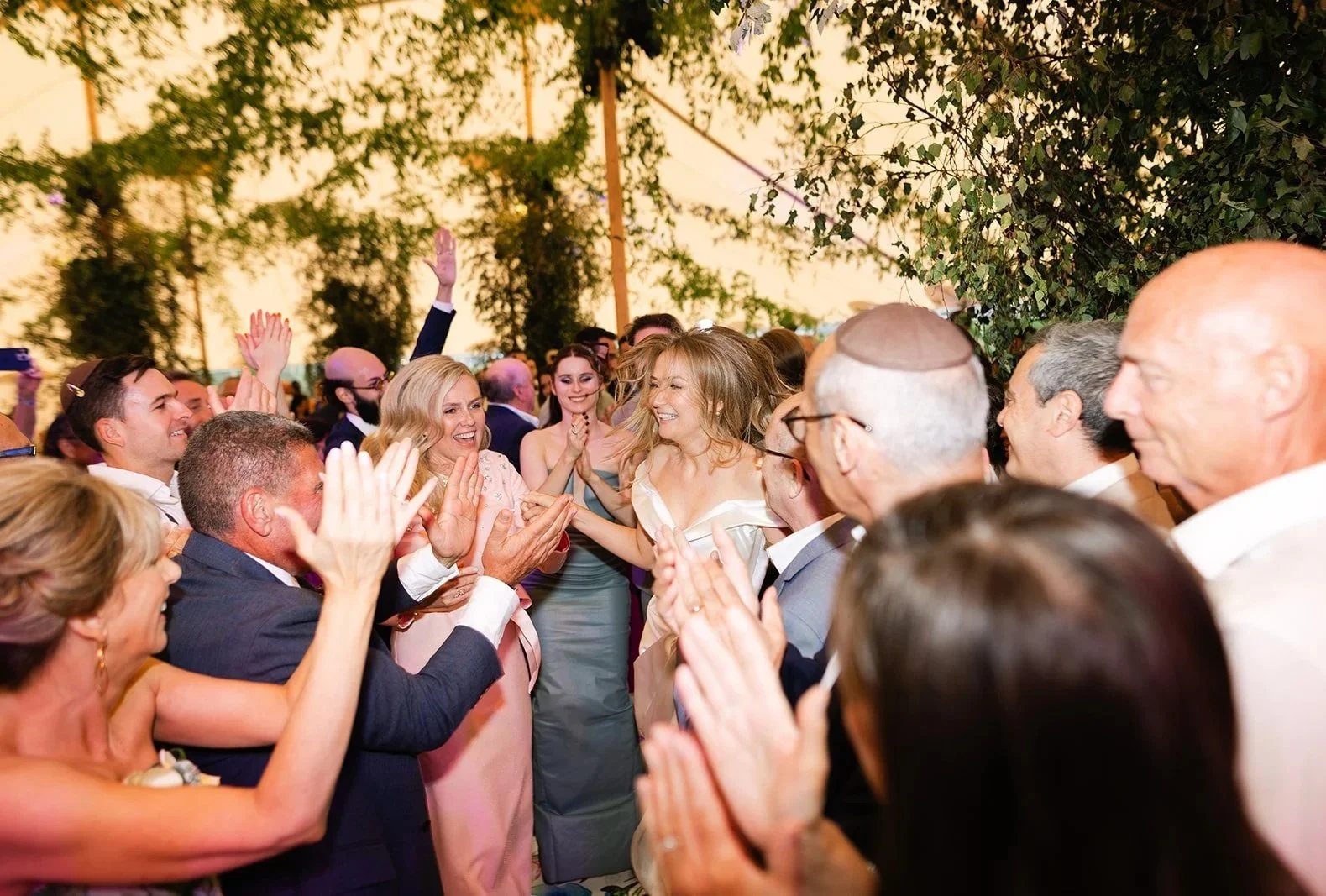 Celebration at a wedding reception with guests clapping, smiling, and dancing under a tent with trees in the background.