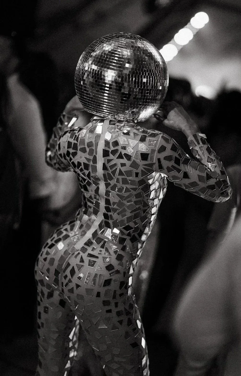 A figure with a reflective, mosaic-patterned body and a disco ball head, viewed from behind, in a dark setting with blurred lights in the background.