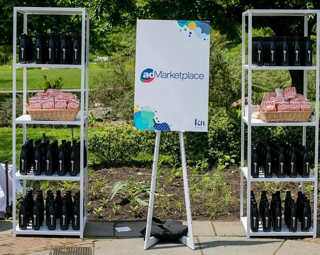 Two white shelves filled with black water bottles and a basket of red and white packaged snacks, with a sign for 'adMarketplace' in the middle, outdoors in a garden area.
