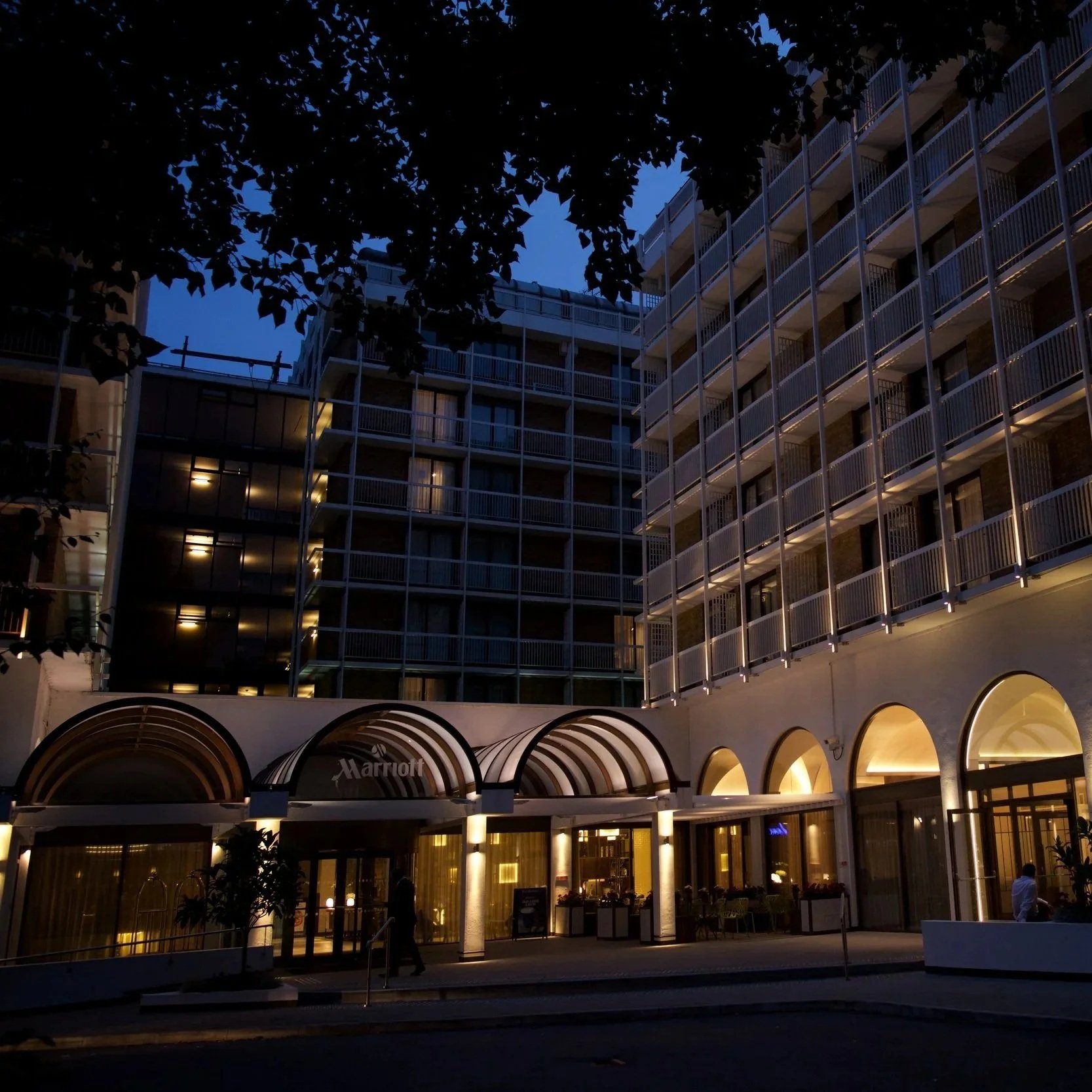 Nighttime view of a multi-story hotel with illuminated ground floor entrance, arched windows, and balconies, with trees partially covering the building.