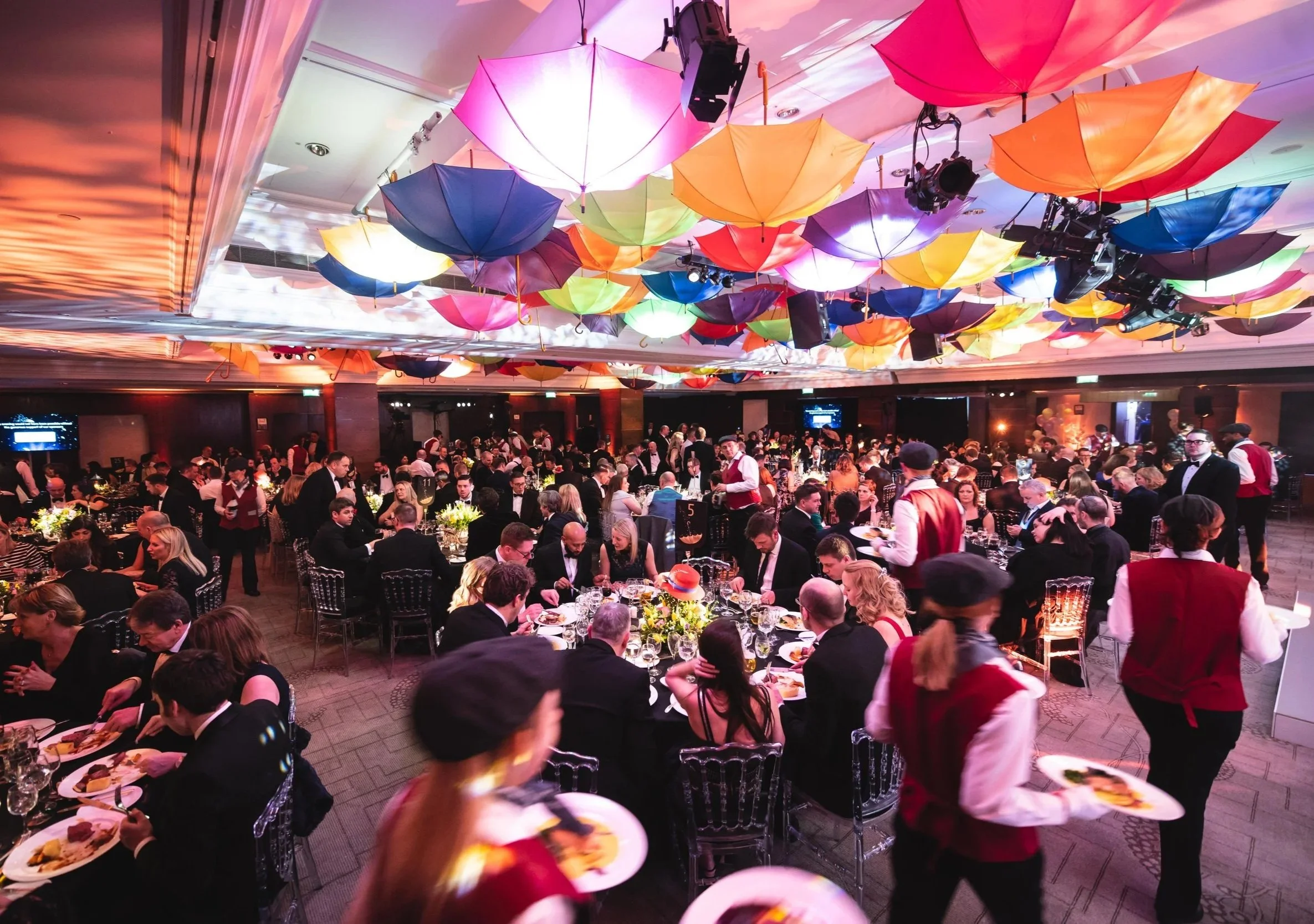 A crowded banquet hall with many guests seated at tables decorated with flowers. Colorful umbrellas are hanging from the ceiling, creating a festive atmosphere.