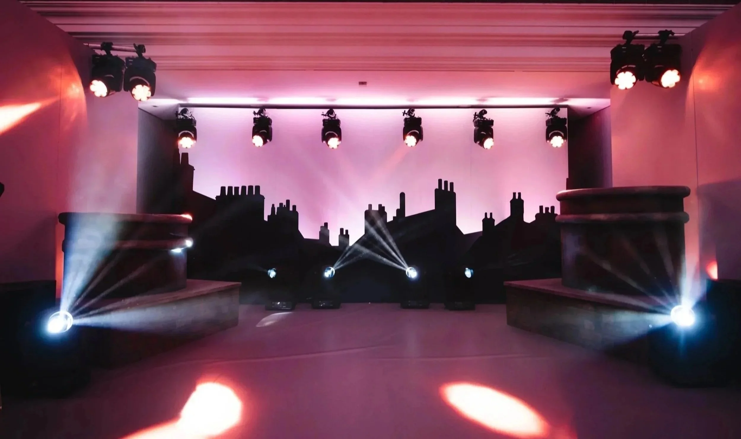 Stage with black city skyline silhouette backdrop, mounted lights on ceiling, and colored spotlights on the floor creating beams of lights.