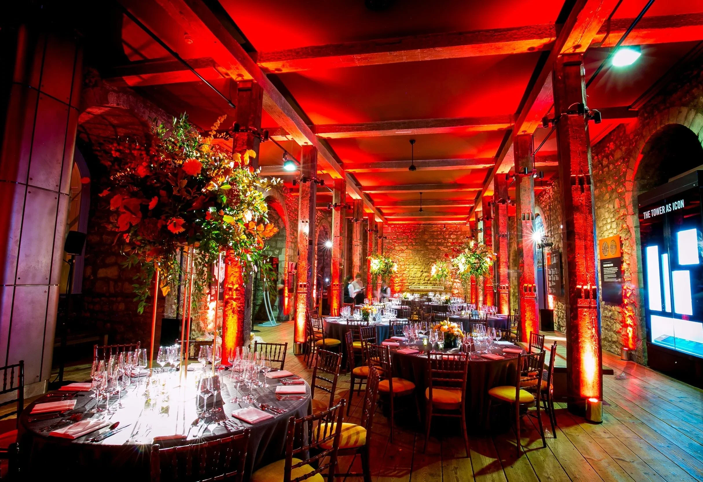 At the Tower of London brick walls, wooden beams, and dramatic red lighting. Round tables are set with glassware, napkins, and silverware, and decorated with floral centerpieces.