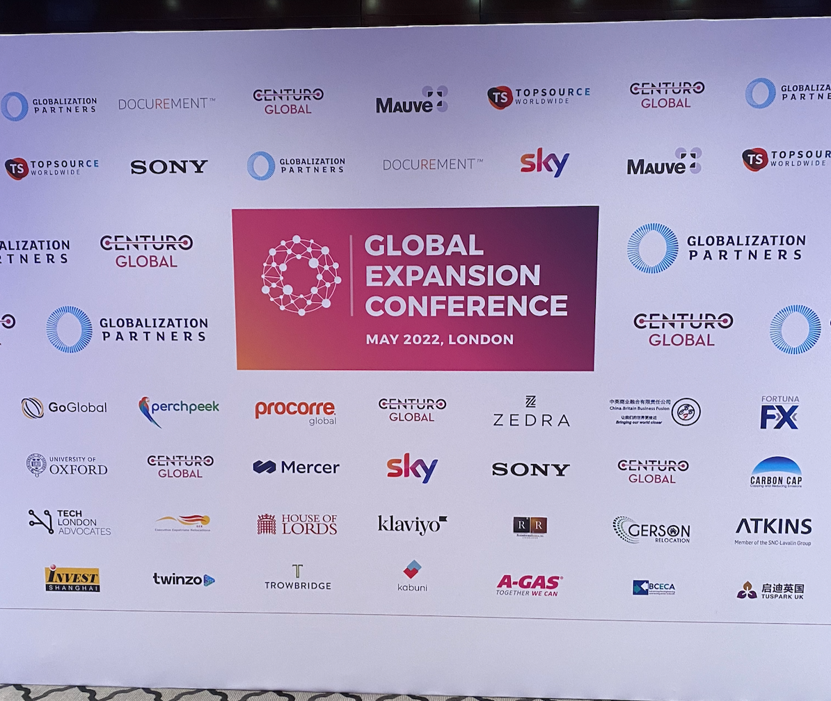 Signboard with logos of various companies, announcing the 'Global Expansion Conference' held in May 2022 in London, with a central graphic of interconnected nodes.