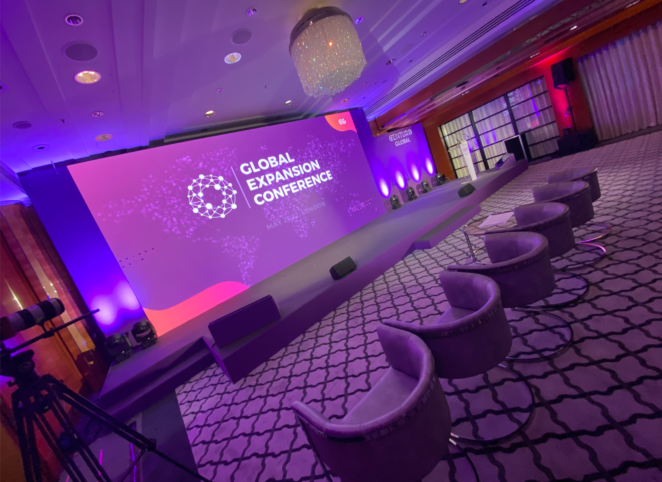 An event stage at the Global Expansion Conference in London, May 2022, with a large purple screen displaying event information, surrounded by chairs, lighting, and a chandelier in a conference room.