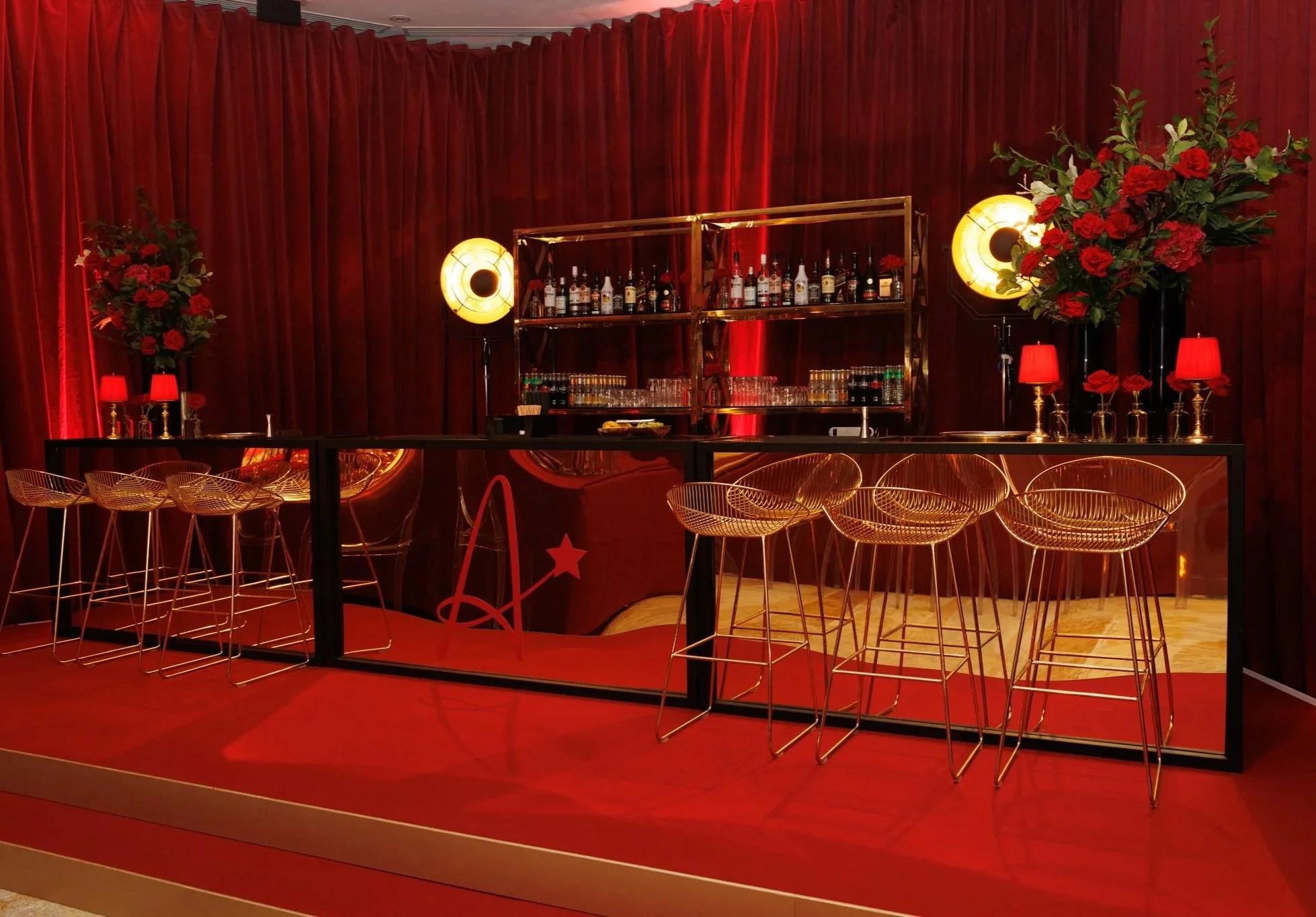 A red-themed bar area with a black counter, gold shelves holding various bottles of alcohol, and decorative red flowers and lamps. There are gold wire chairs in front of the bar.