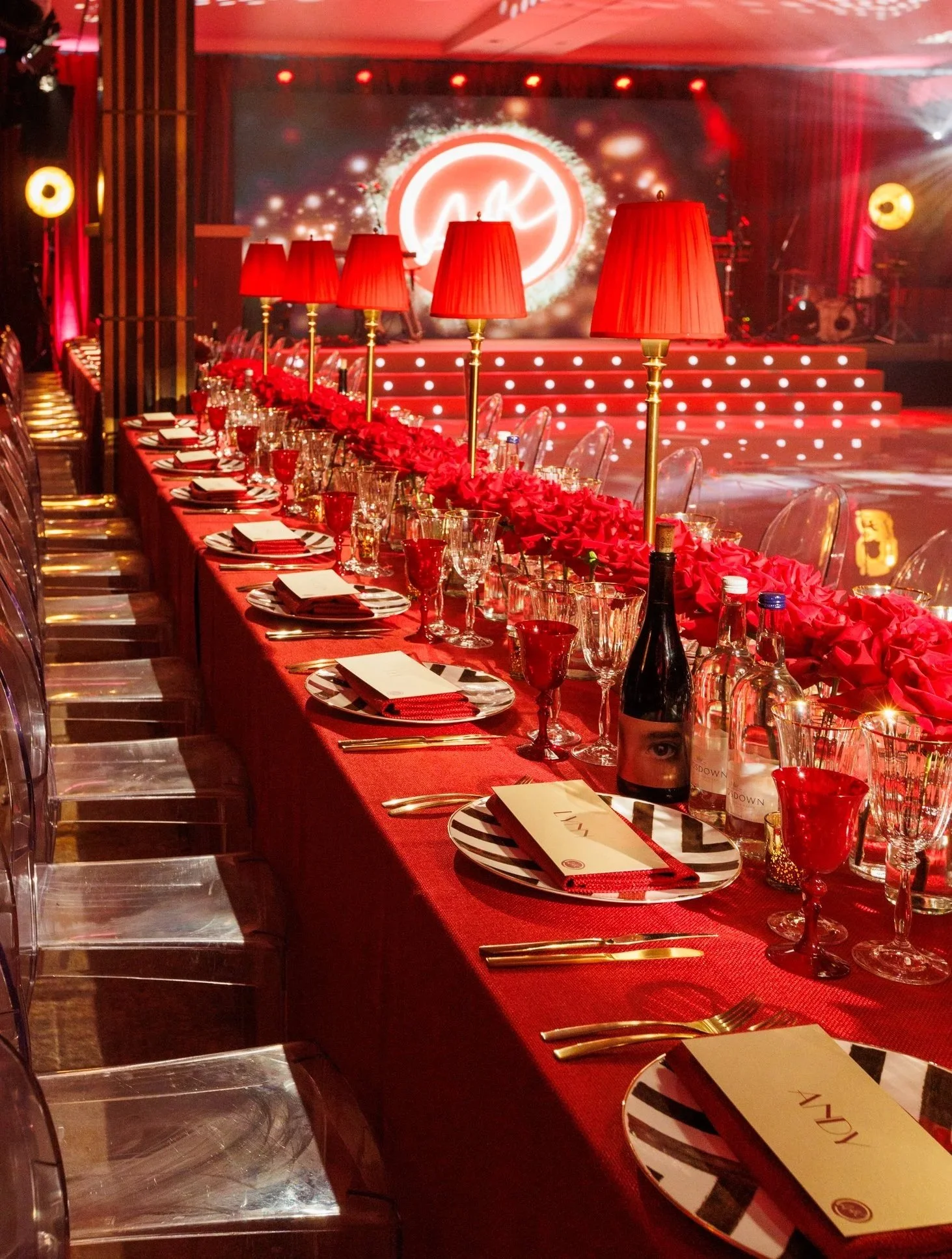 A decorated dinner table set for a celebration or event with a red tablecloth, festive black and white striped plates, red cups, and gold utensils. Red lamps with gold stands are evenly spaced along the table, and a long floral centerpiece with red f