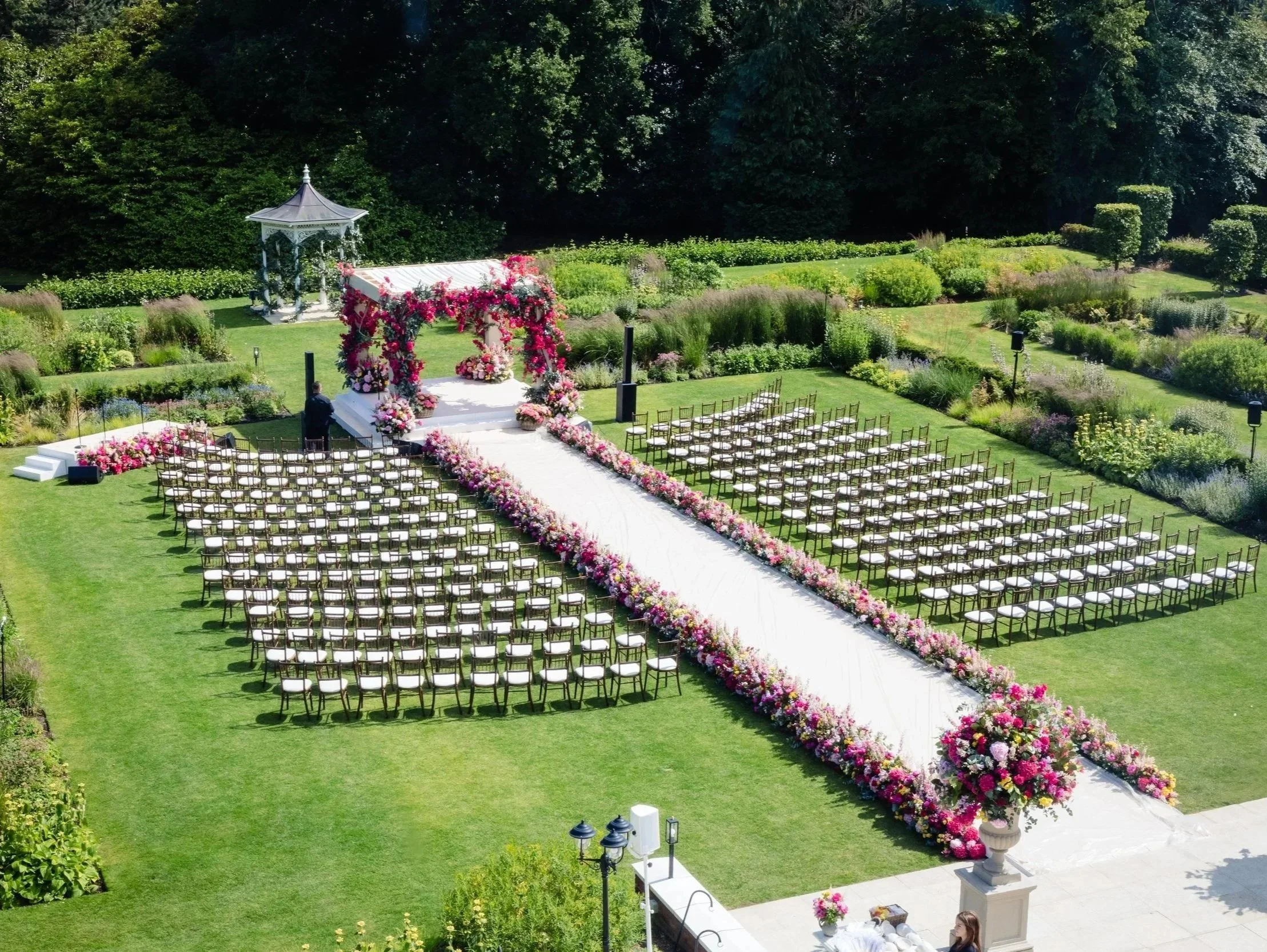 An outdoor wedding ceremony setup with rows of white chairs and a white aisle skirted with pink and purple flowers. At the front, there is a floral arch with pink and purple flowers and a gazebo-like structure behind it, all set in a garden with lush