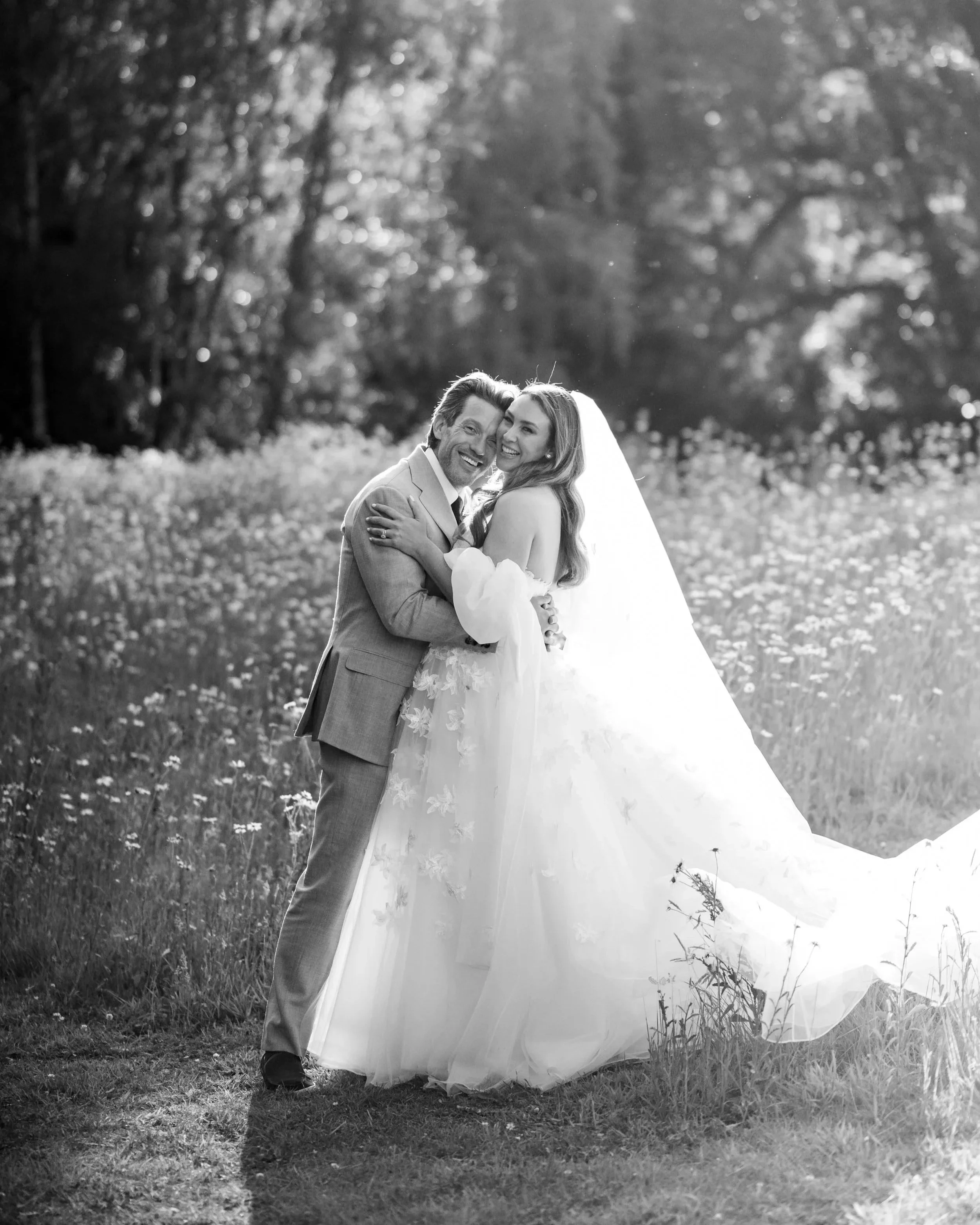 Black-and-white photo of a newlywed couple standing outdoors on grass, smiling and hugging. The groom is wearing a suit, and the bride is in a wedding dress with a veil. Behind them are blurred trees and sunlight filtering through the leaves.