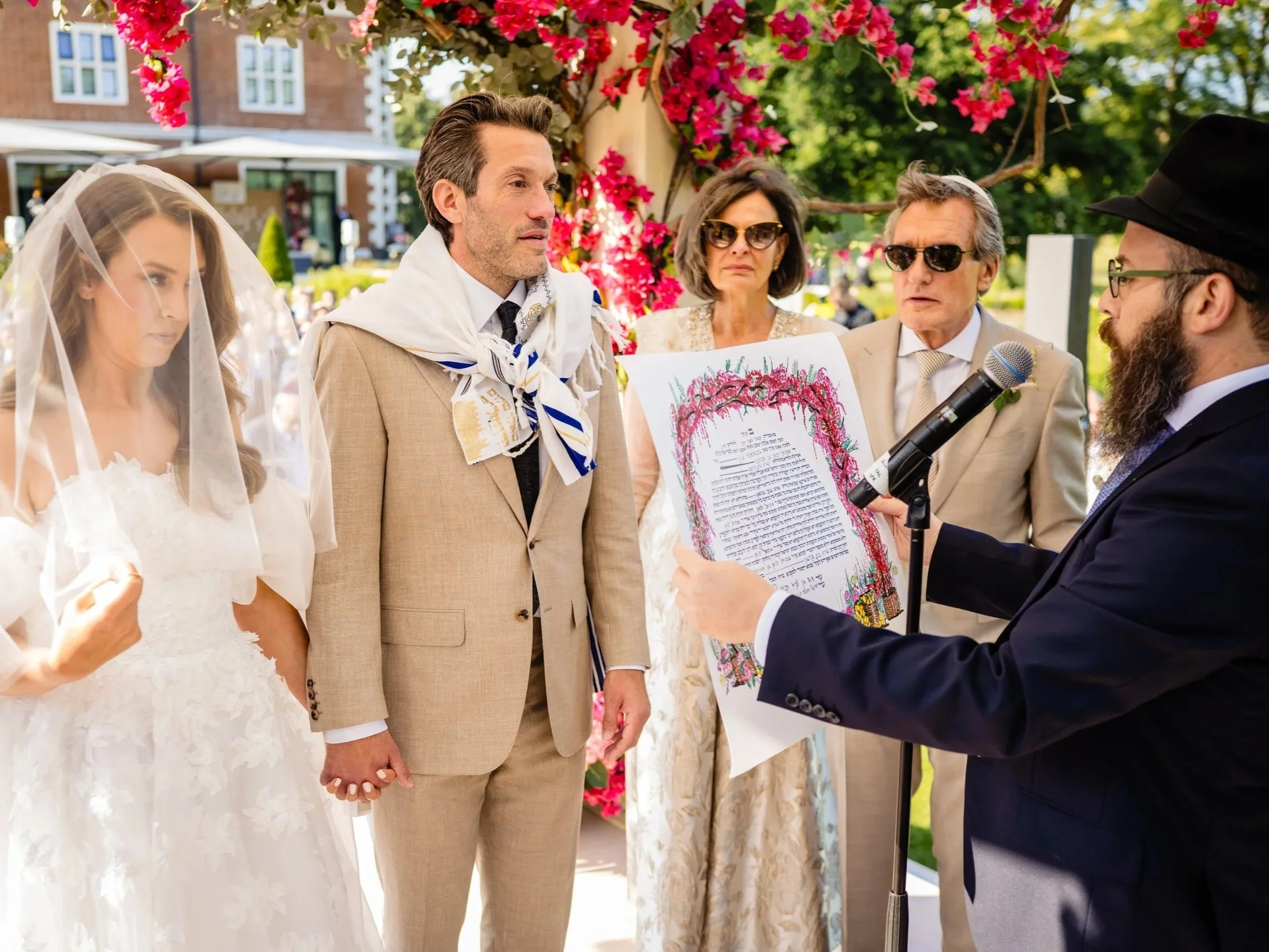 A wedding ceremony outdoors with the bride, groom, and officiant holding hands amidst friends and family. The bride is wearing a white dress with a veil, and the groom is wearing a beige suit and tie. The officiant, holding a microphone, is reading f