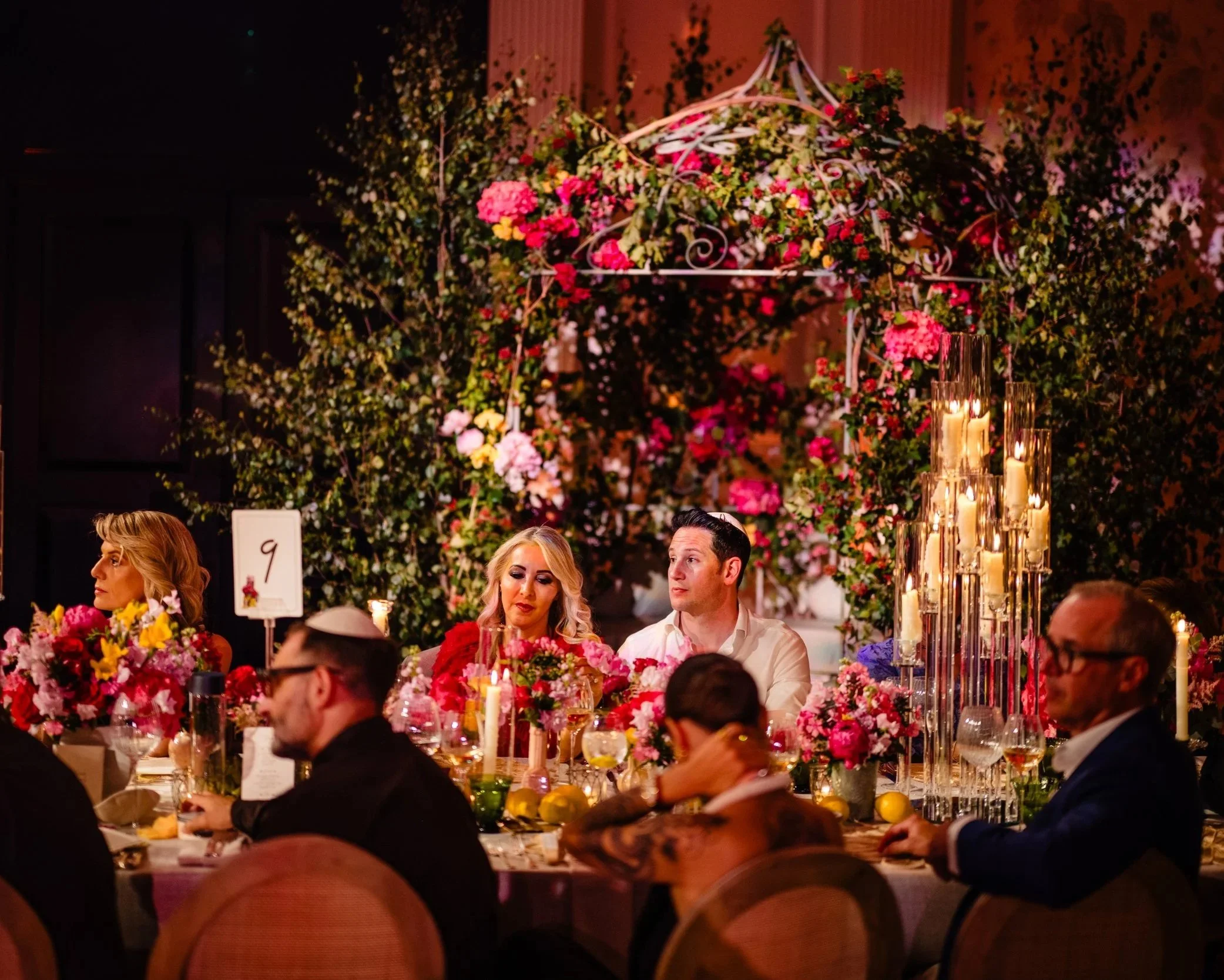 A group of people sitting around a table decorated with pink flowers, candles, and glasses at a banquet or wedding reception inside a venue with a floral overhead decoration and lit candles at Fairmont Windsor Park